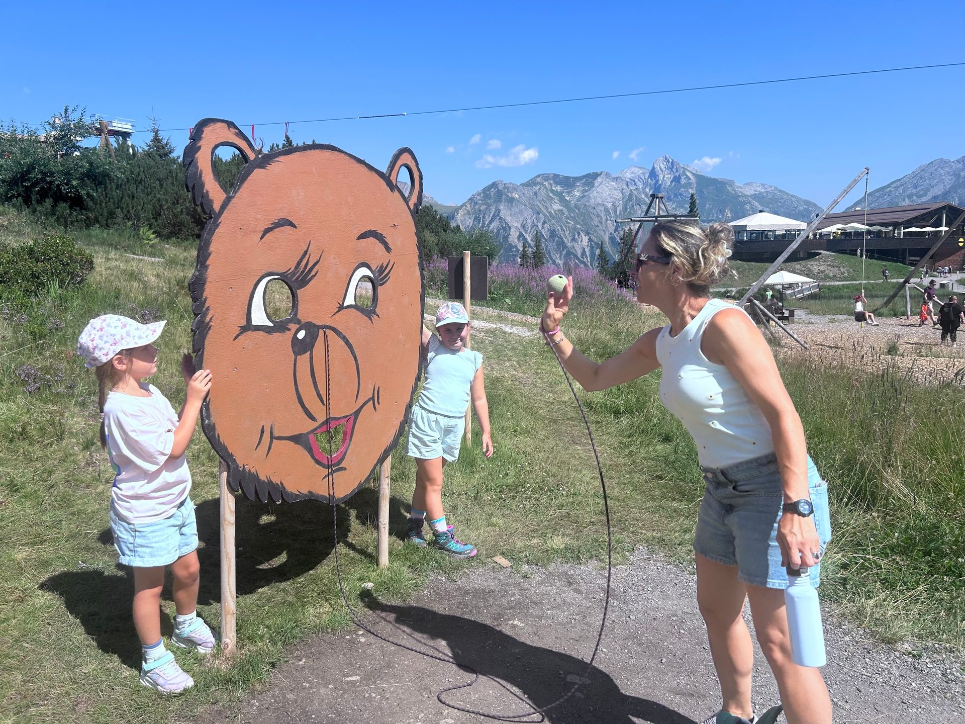 A woman and two girls play with a bear-shaped cutout target in an alpine setting on a sunny day.