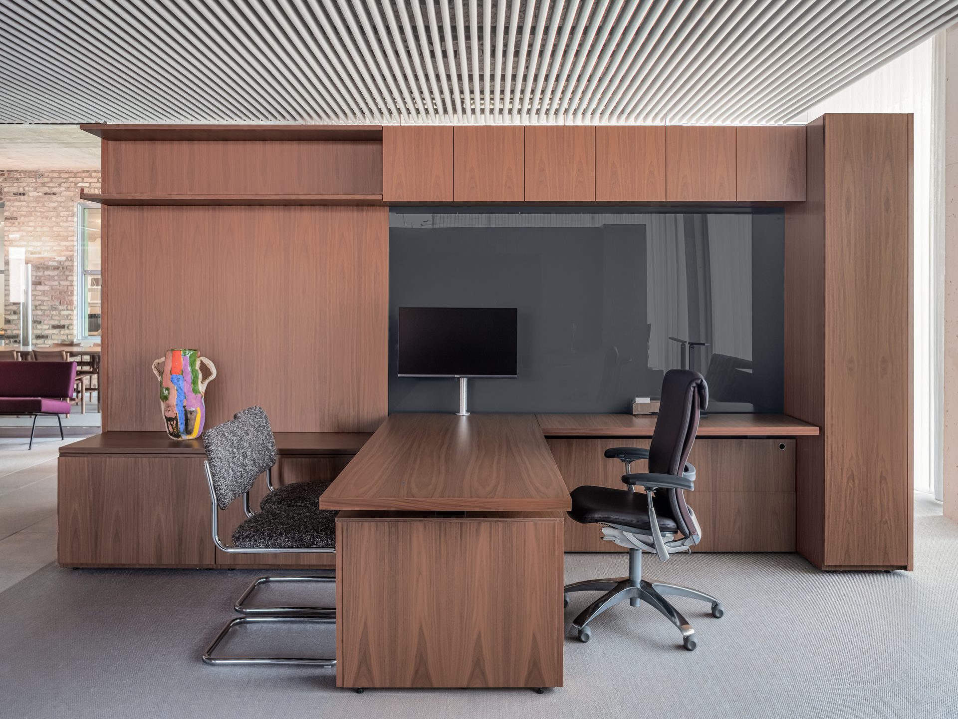 Contemporary office with dark wood desk, storage, chairs, monitor, colorful vase, slatted ceiling.