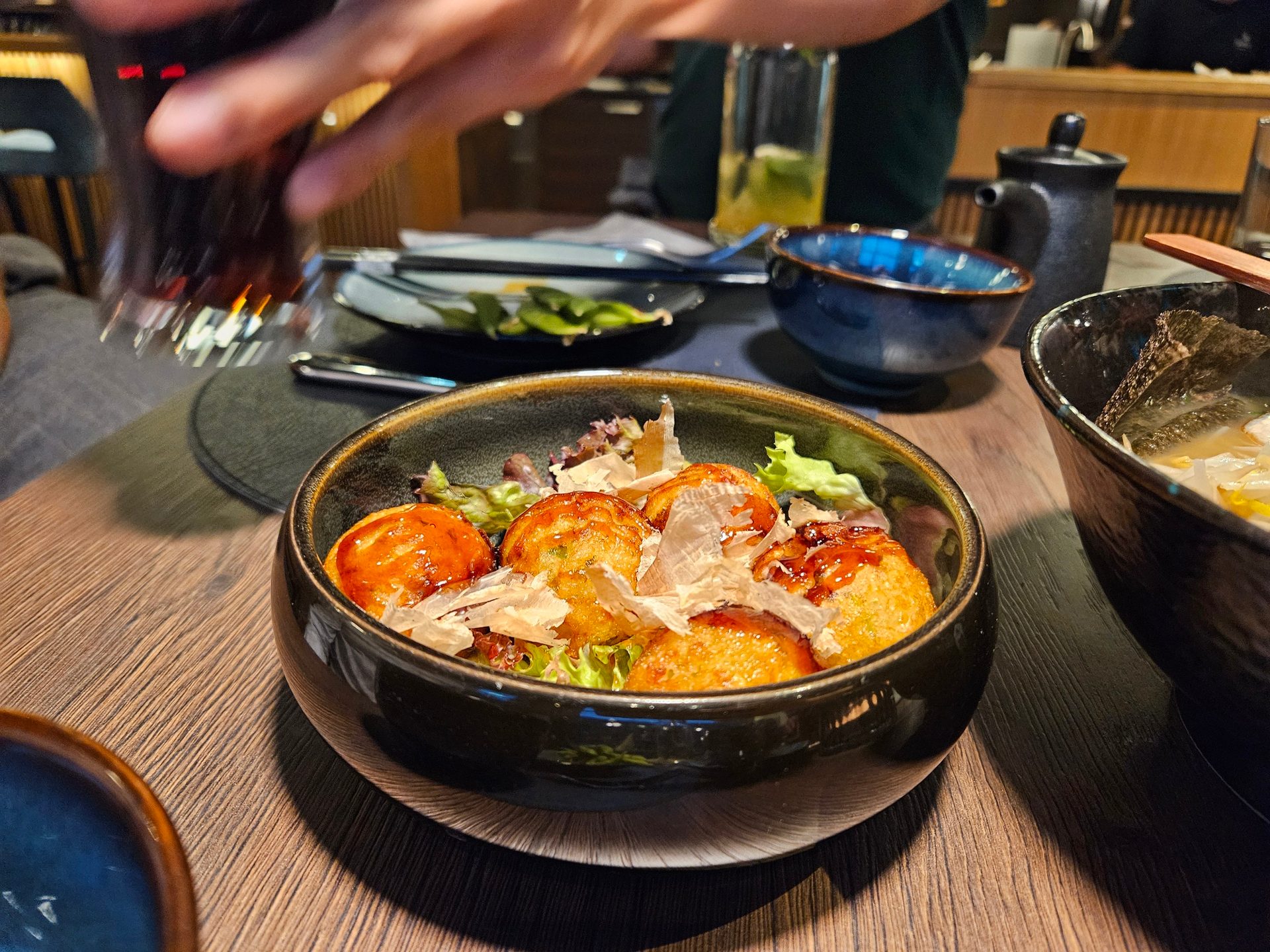 A bowl of takoyaki with bonito flakes and sauce on a wooden table in a dimly lit restaurant.