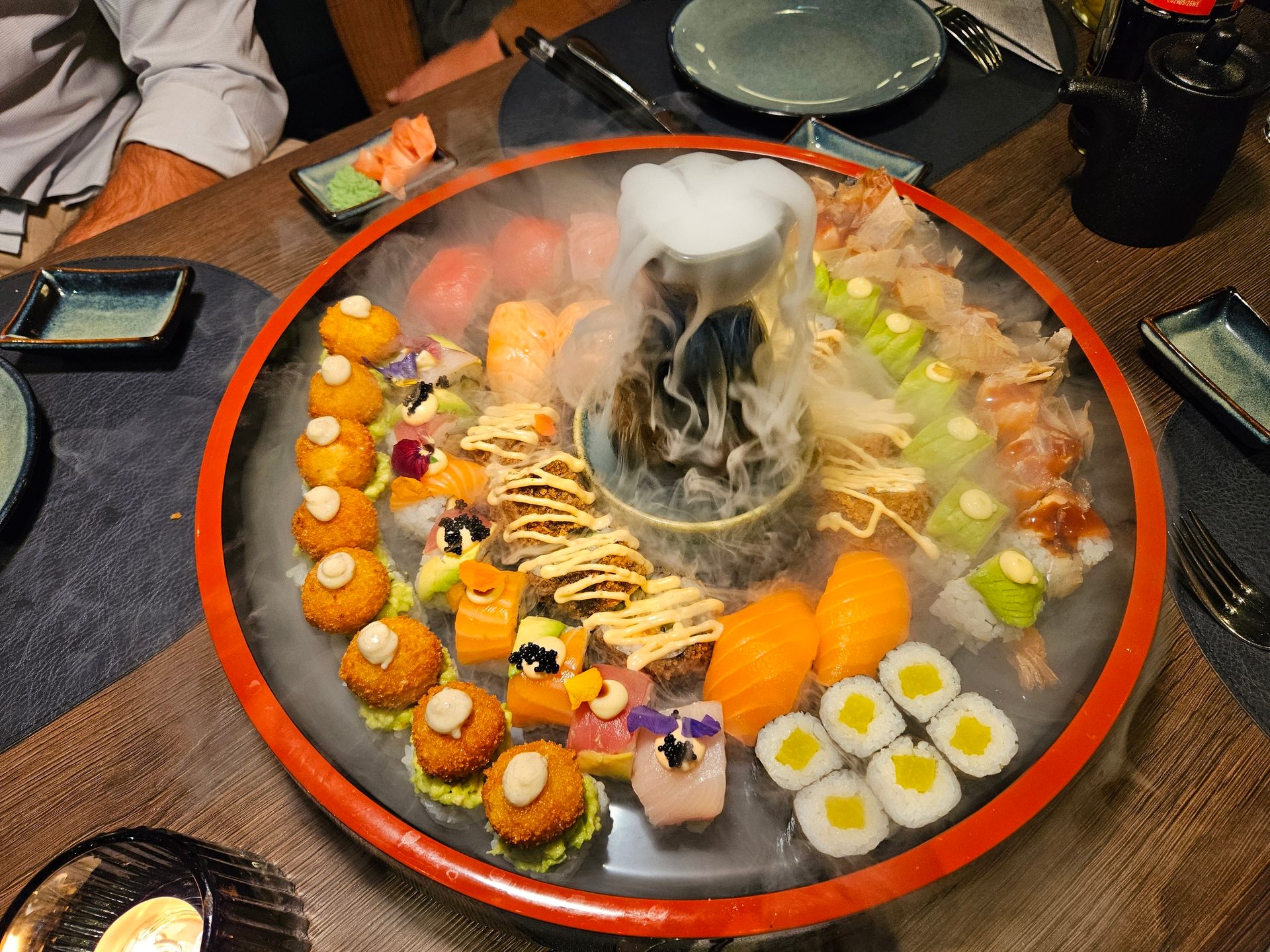 Large sushi platter with dry ice smoke, featuring a variety of nigiri, rolls, and fried sushi.