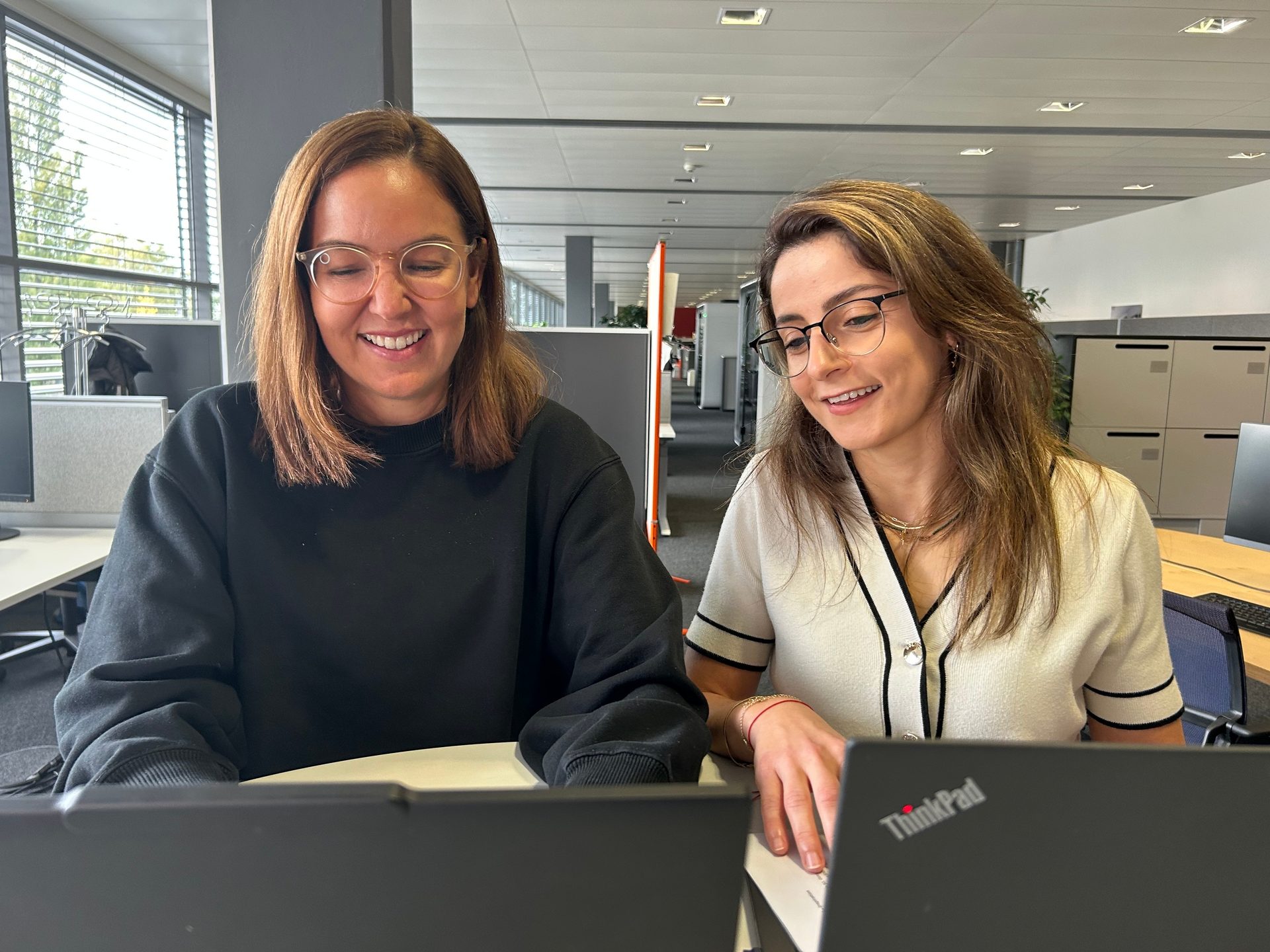 Two smiling women in glasses looking at a laptop in an office.