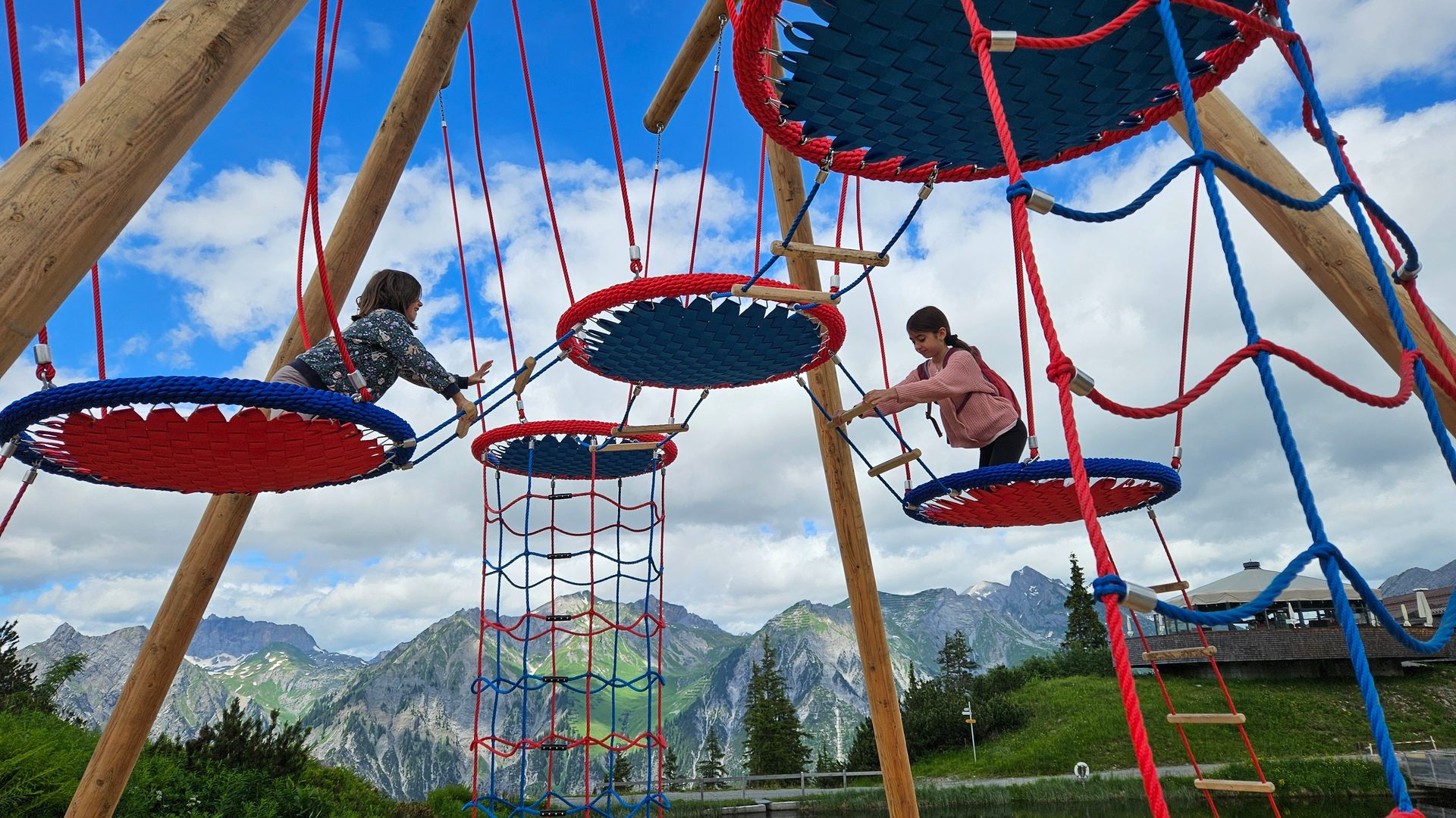 Two girls play on a vibrant ropes course, suspended over a scenic mountain lake with clouds.