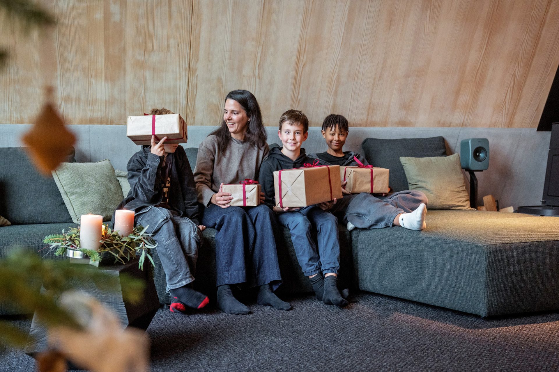 A family of four sits on a sofa with Christmas gifts; one child's face is hidden by a present.