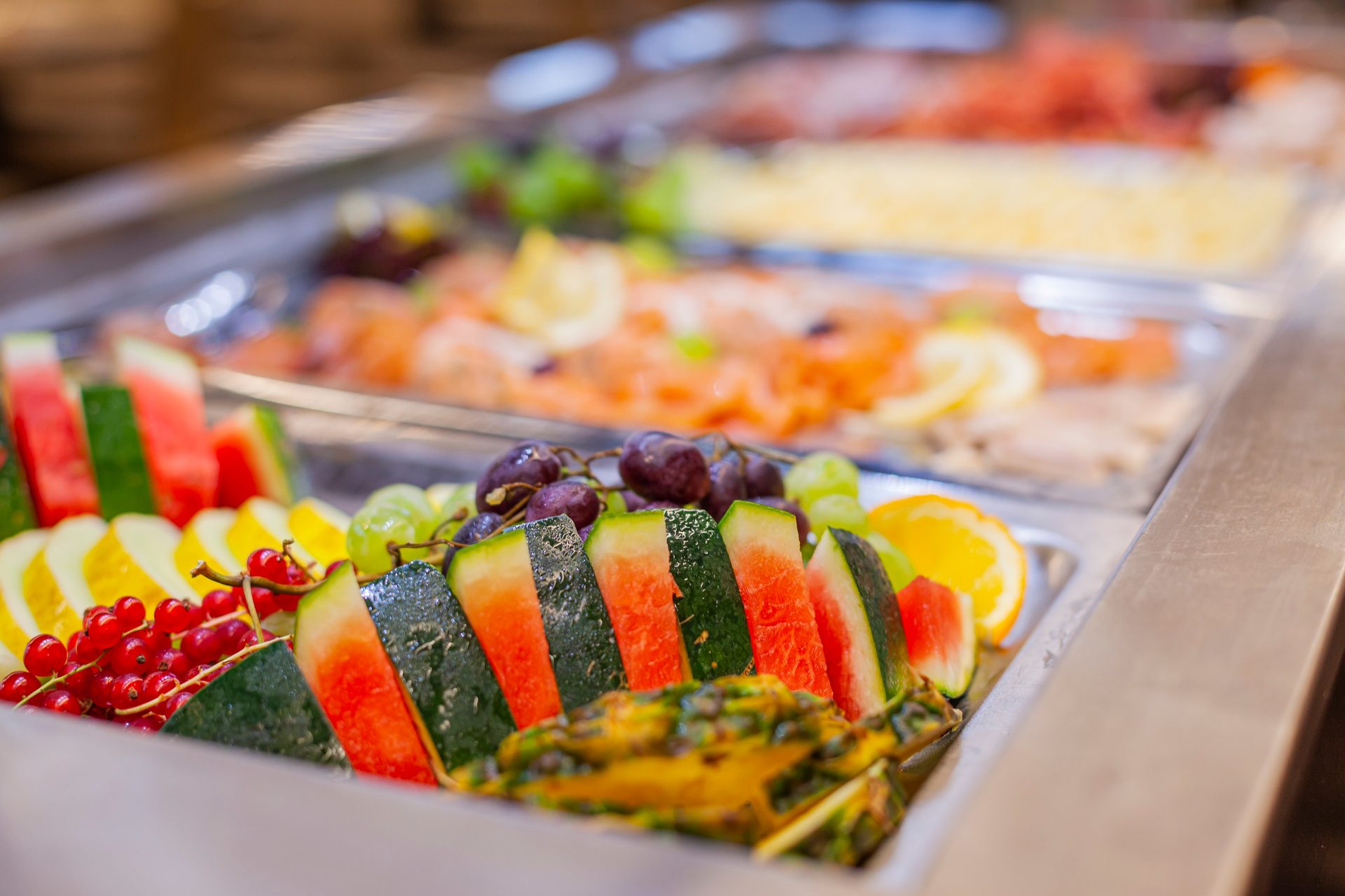 Vibrant buffet with fresh fruit: watermelon, melon, grapes, red currants, and salmon in background.