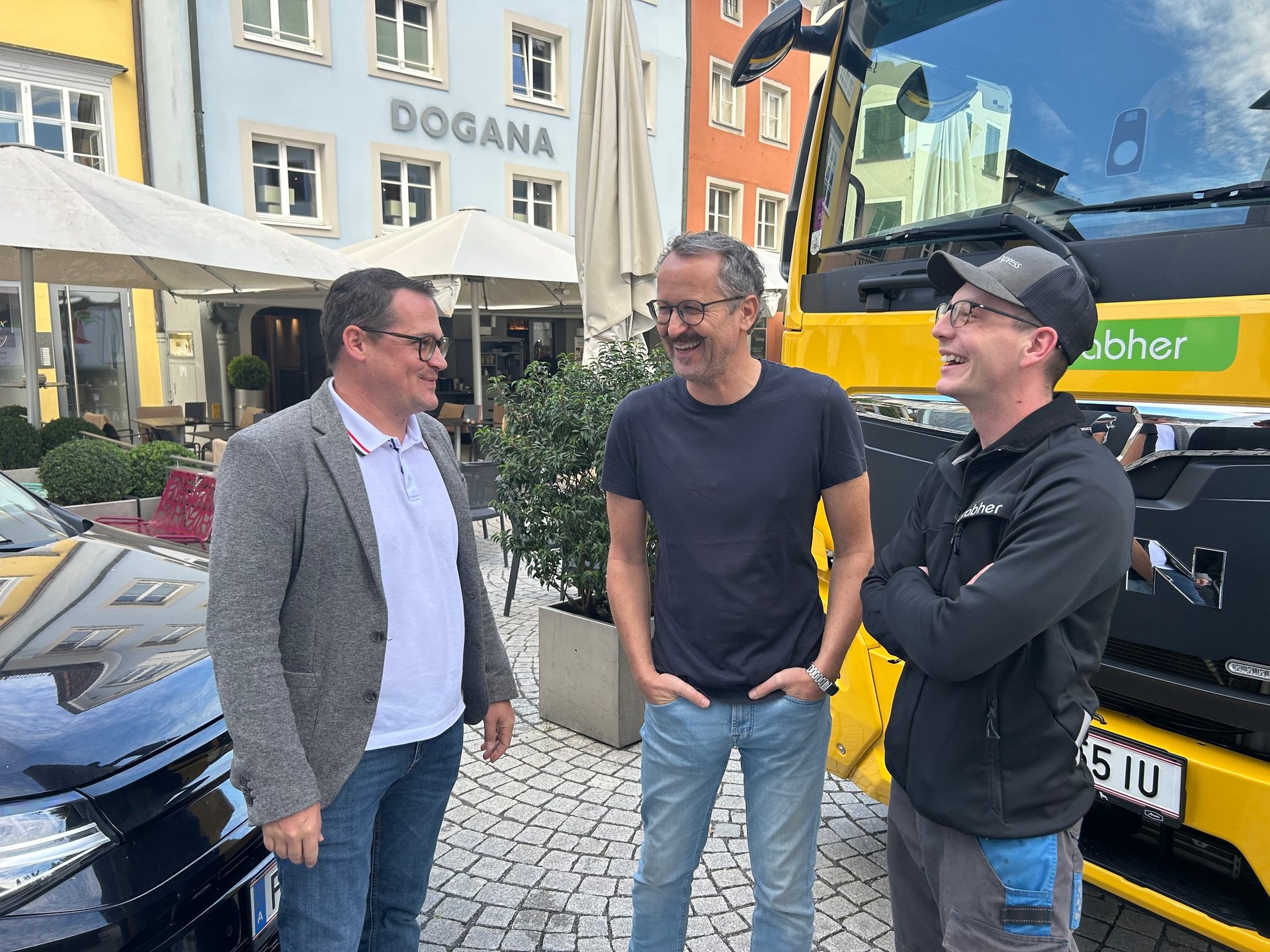 Three men are laughing and talking outdoors, with a yellow truck and a building in the background.