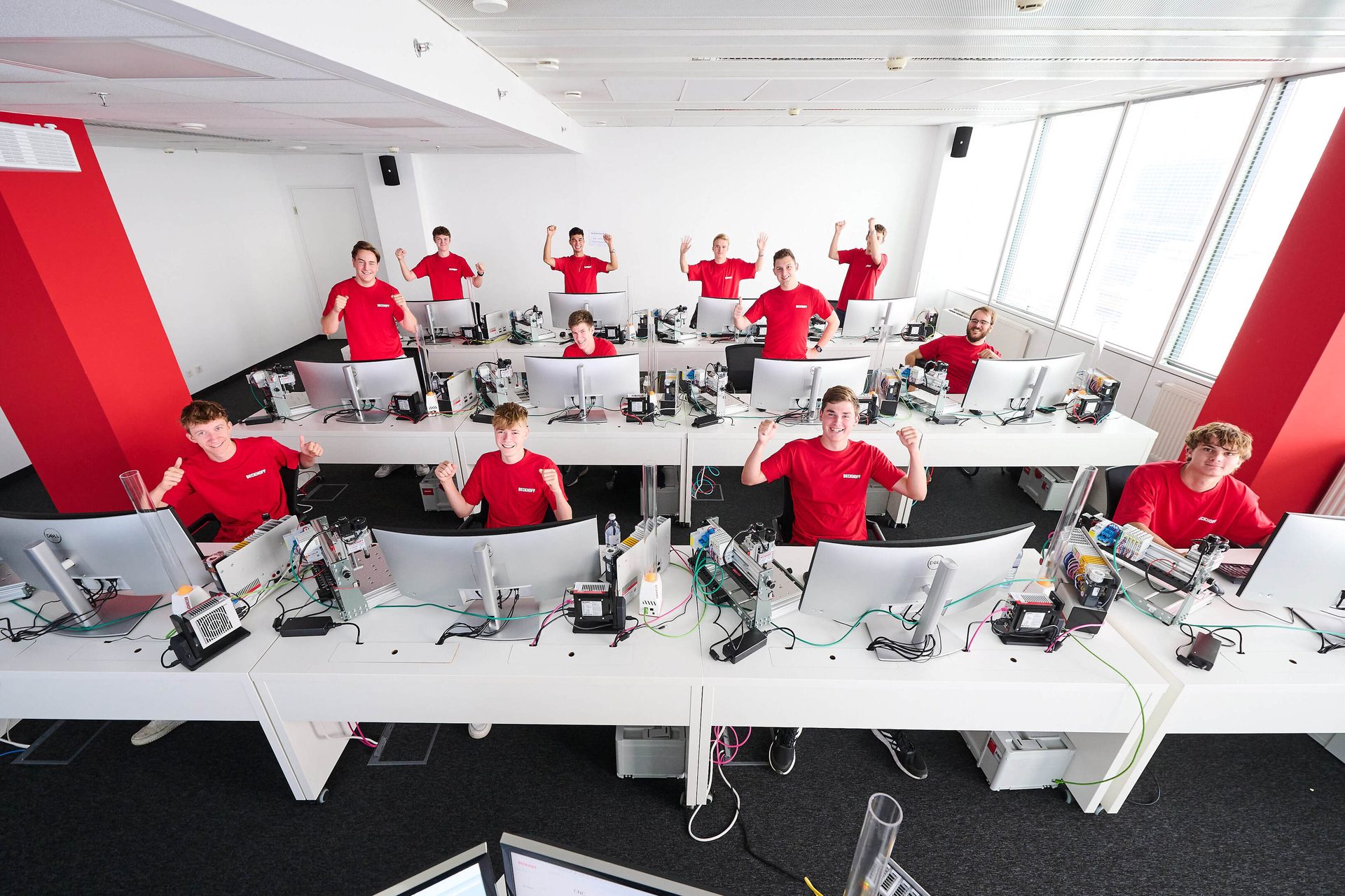 A group of young men in red shirts enthusiastically working at curved computer desks in a modern office.