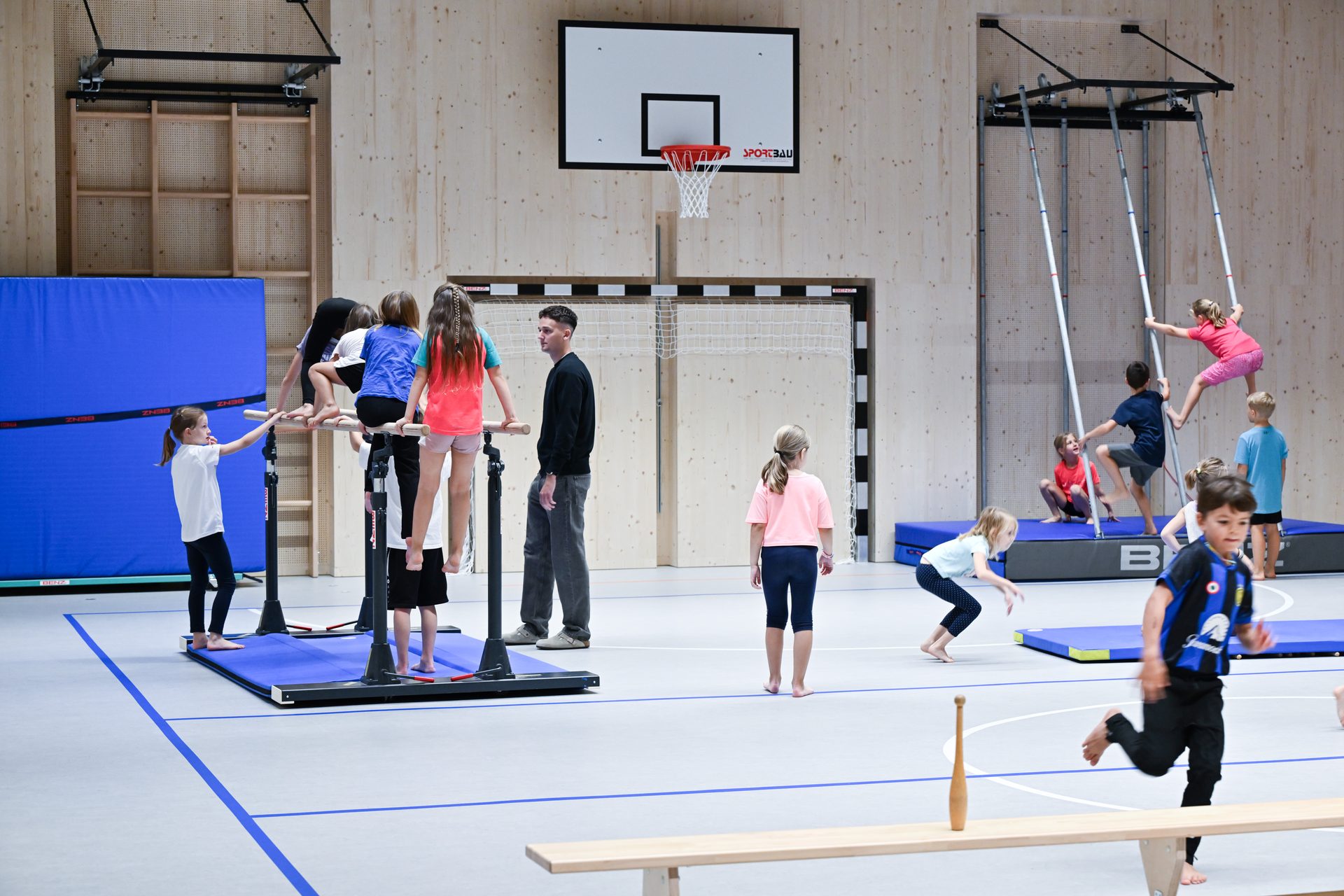 Children and an adult doing gymnastics in a bright gym with wooden walls.