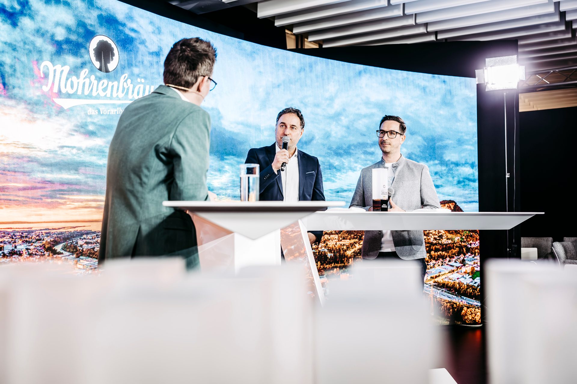 Three men on a stage with a large screen displaying a landscape and brand logo in the background.