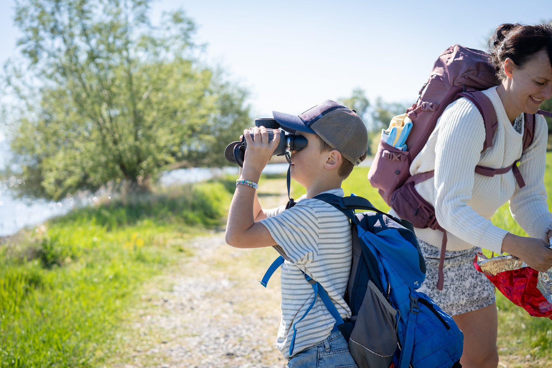 A boy uses binoculars for birdwatching with a woman, both wearing backpacks, on a sunny day.