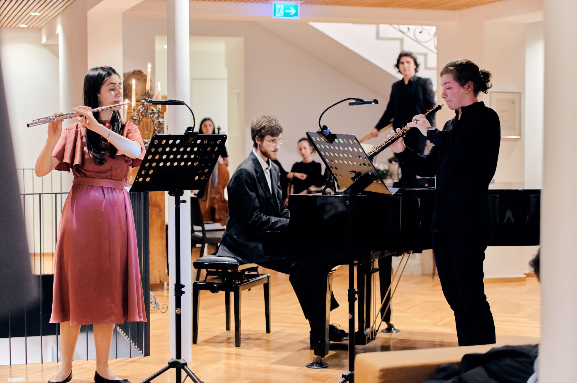 Classical musicians performing: a flutist, pianist, and oboist in the foreground.
