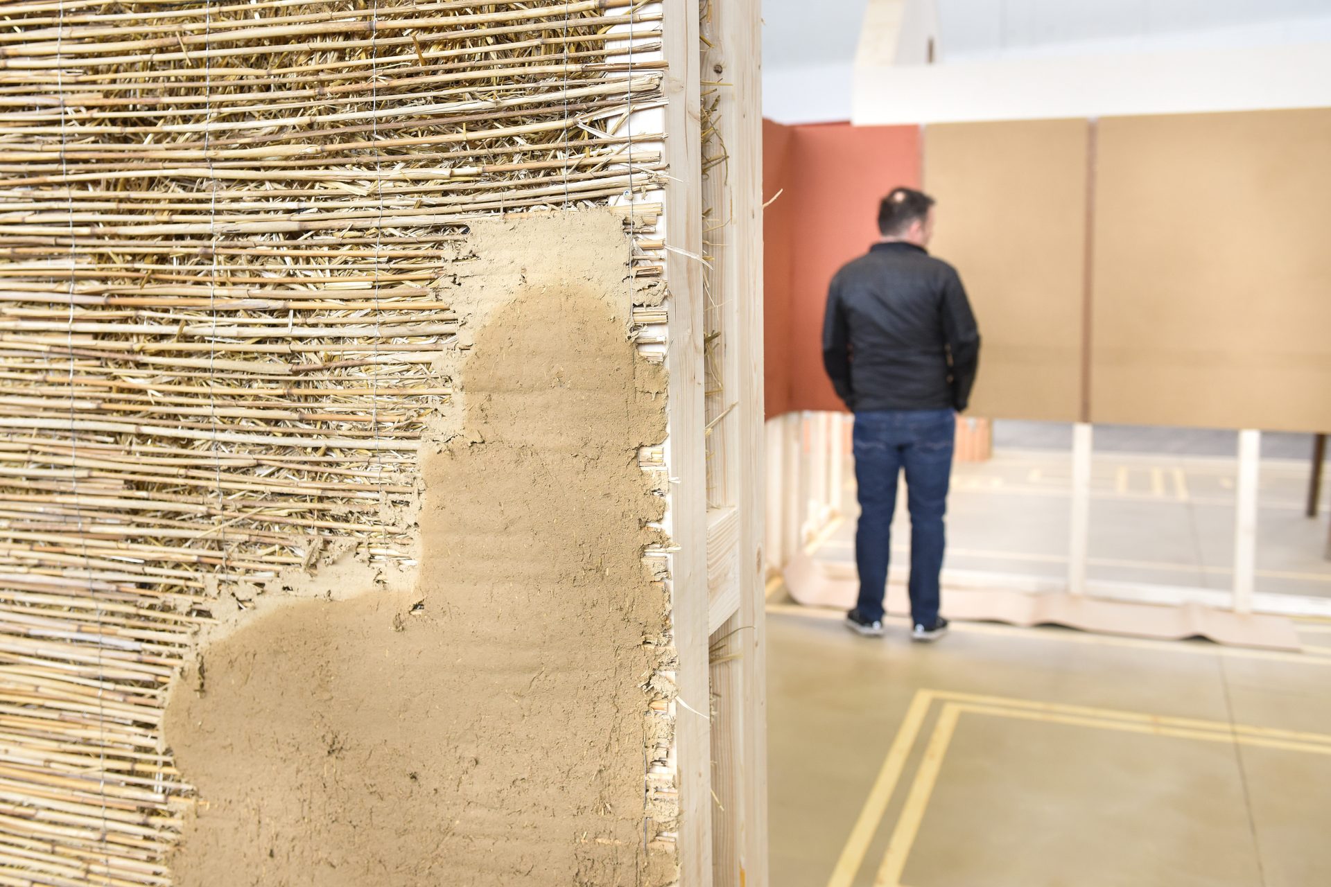 Exposed natural building wall with straw matting and earth plaster; a person views panels in background.