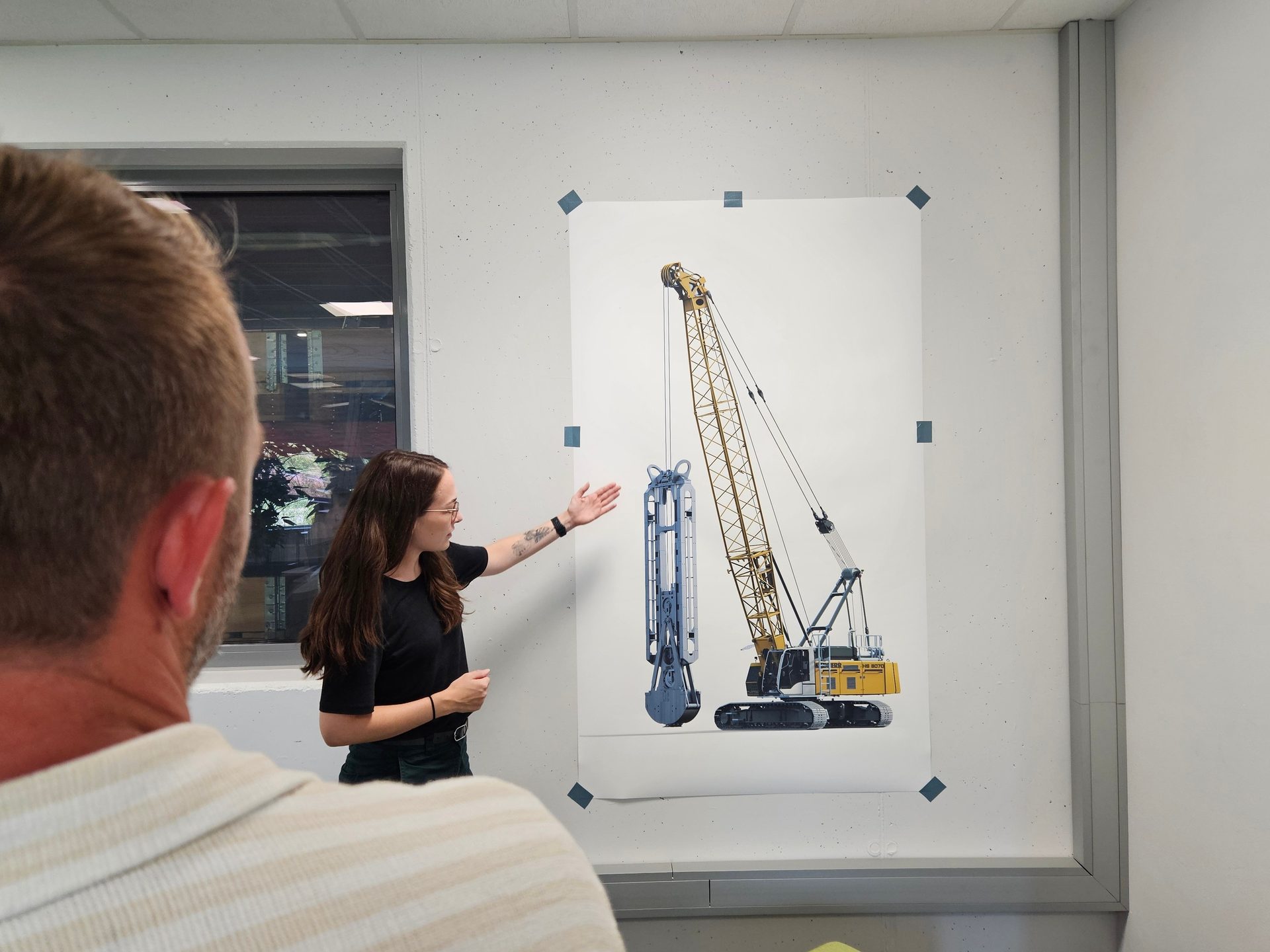 Woman presents a large poster showing a yellow crane with a drilling attachment.