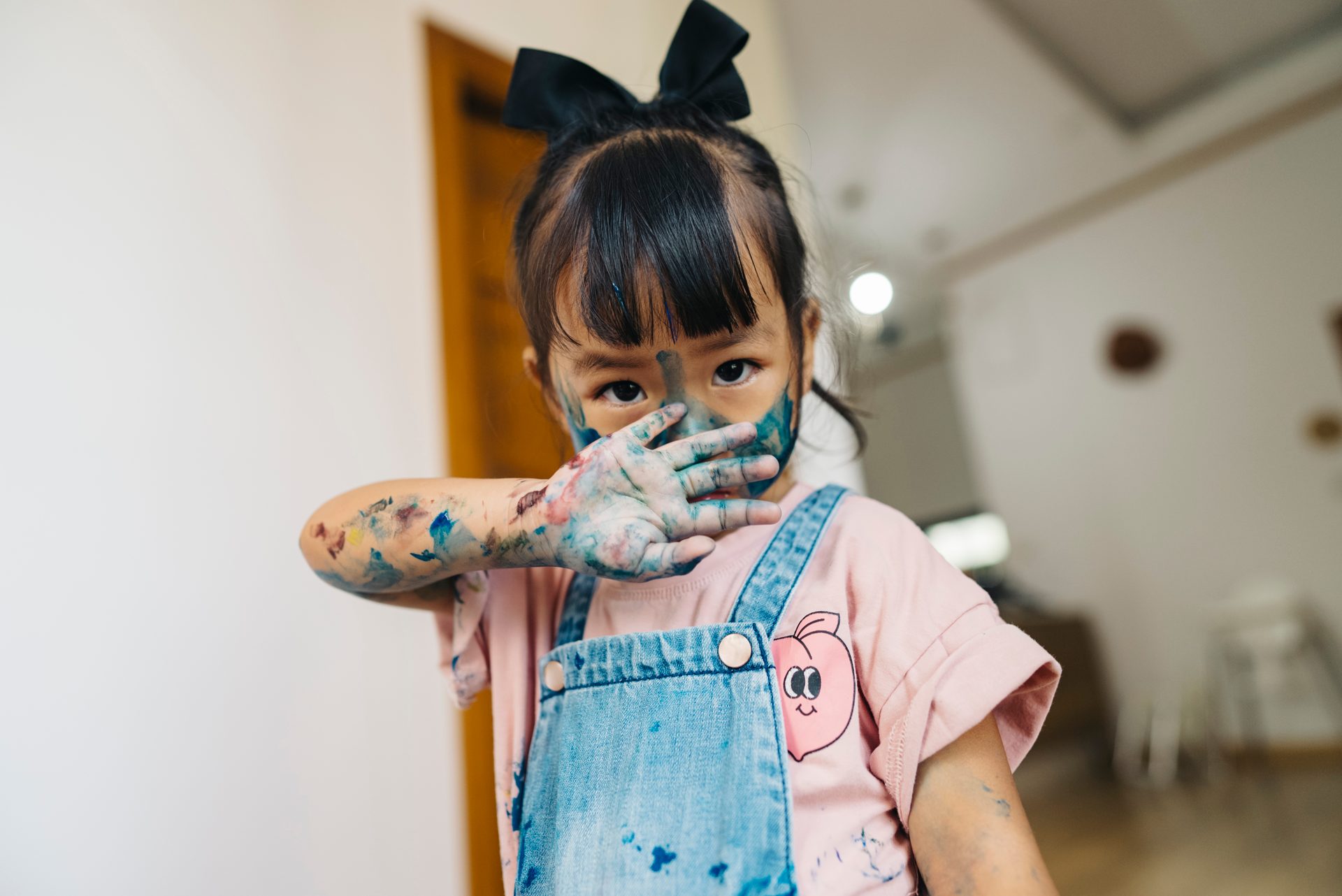 A young child covered in colorful paint, holding a paint-smeared hand over their mouth.