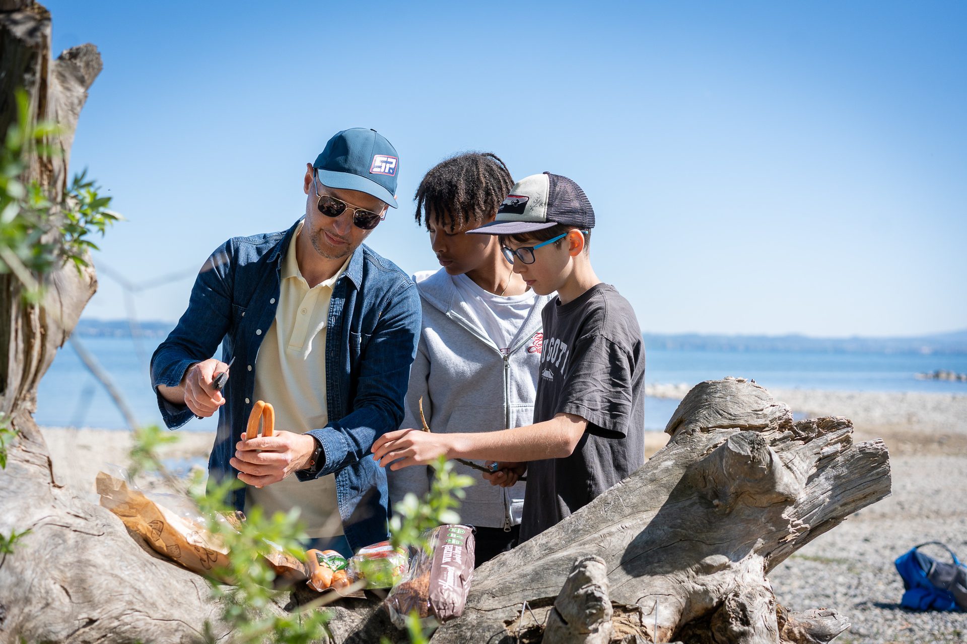 A man and two boys prepare sausages and snacks on a tree trunk by a sunny lake.