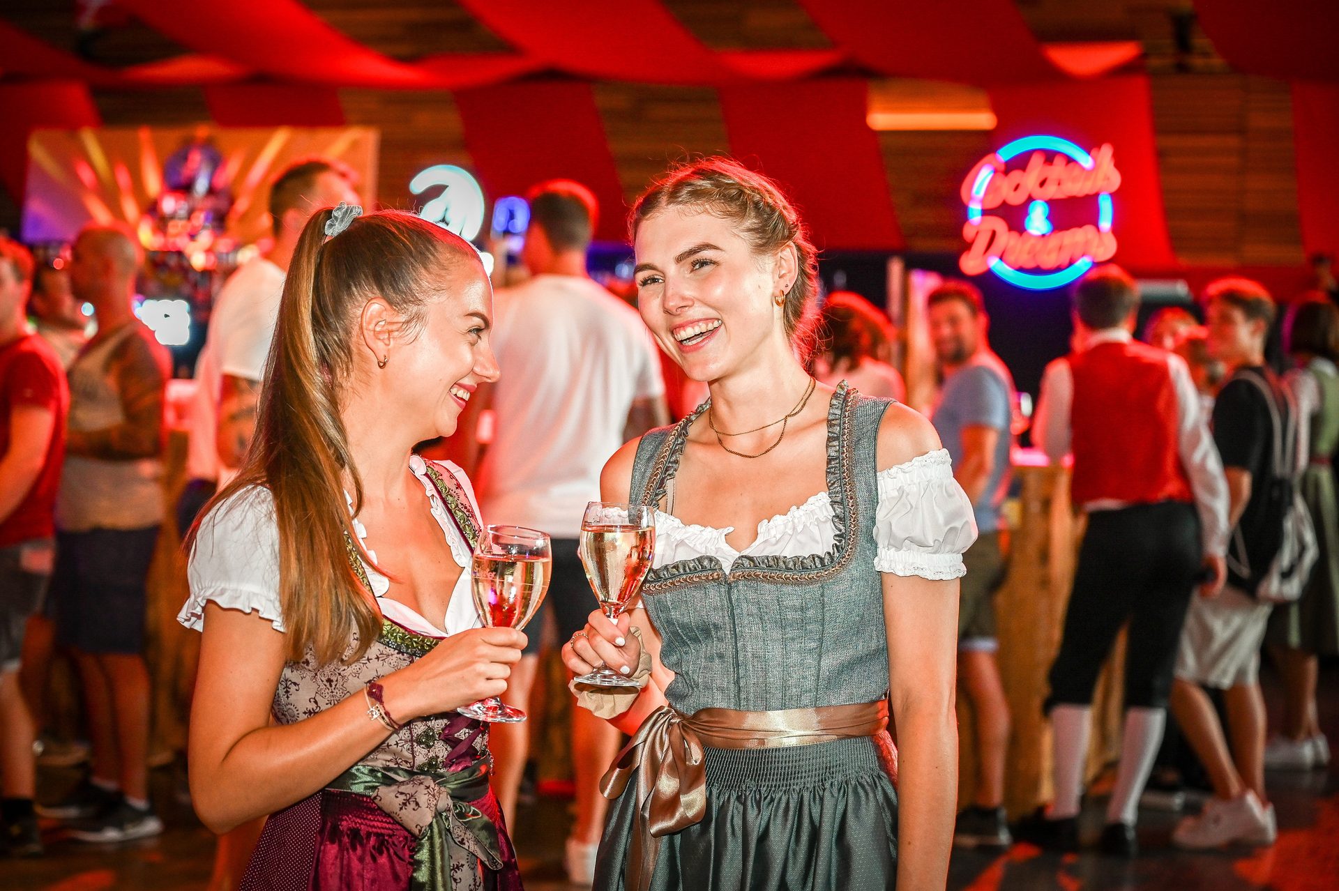 Two smiling women in dirndl dresses clink glasses at a festive event.