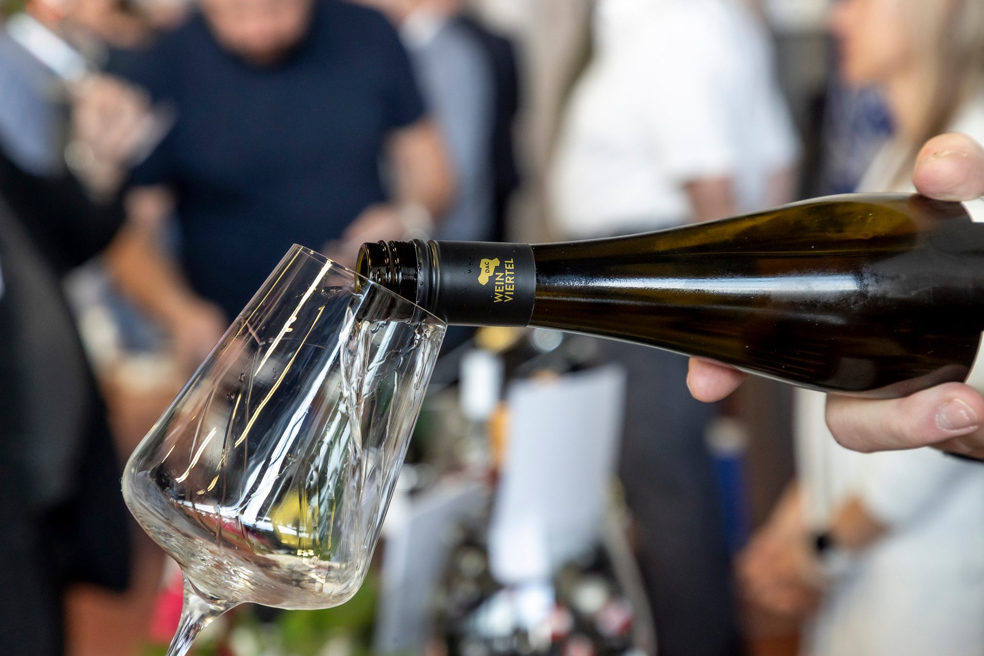Hand pouring white wine from a bottle labeled "DAC Weinviertel" into a clear wine glass at an event.