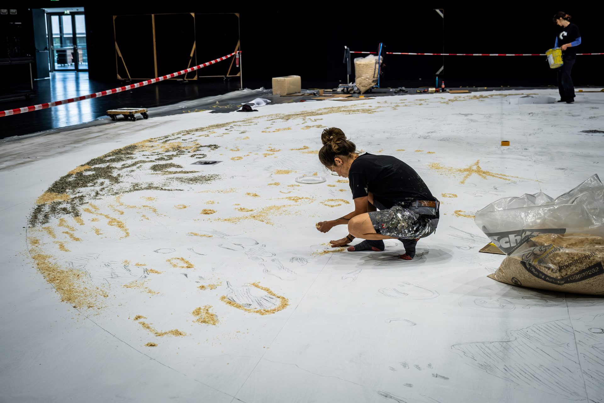 Artist meticulously applies granular material to a large circular floor drawing, creating intricate patterns.