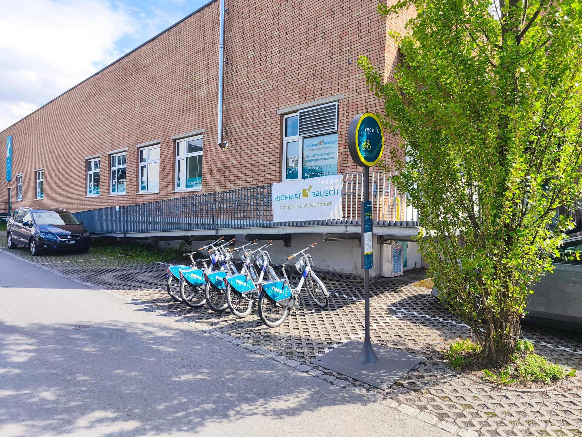 Brick building with rental bikes, a bike share sign, a parked car, and a green tree on a sunny day.