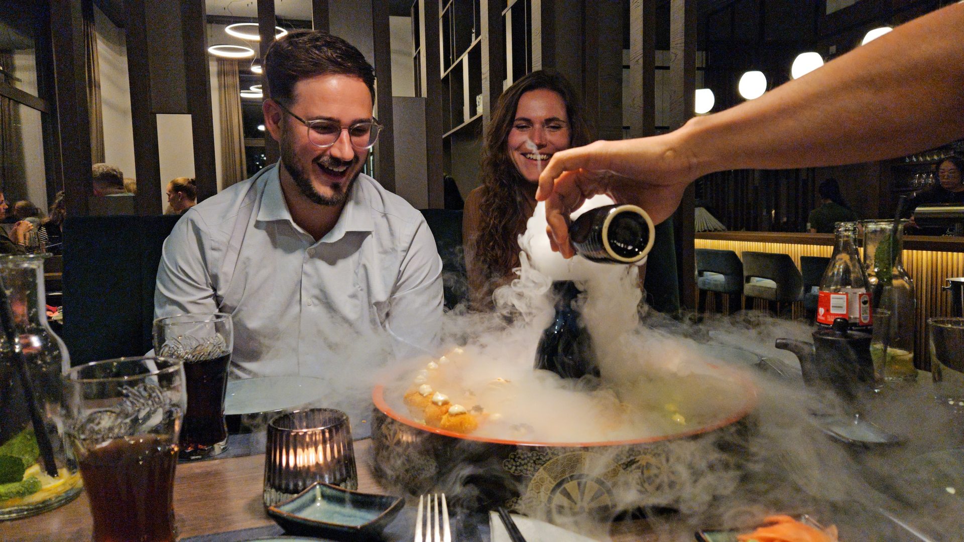 Smiling couple watches a hand pour liquid creating smoke on a dish at a restaurant.