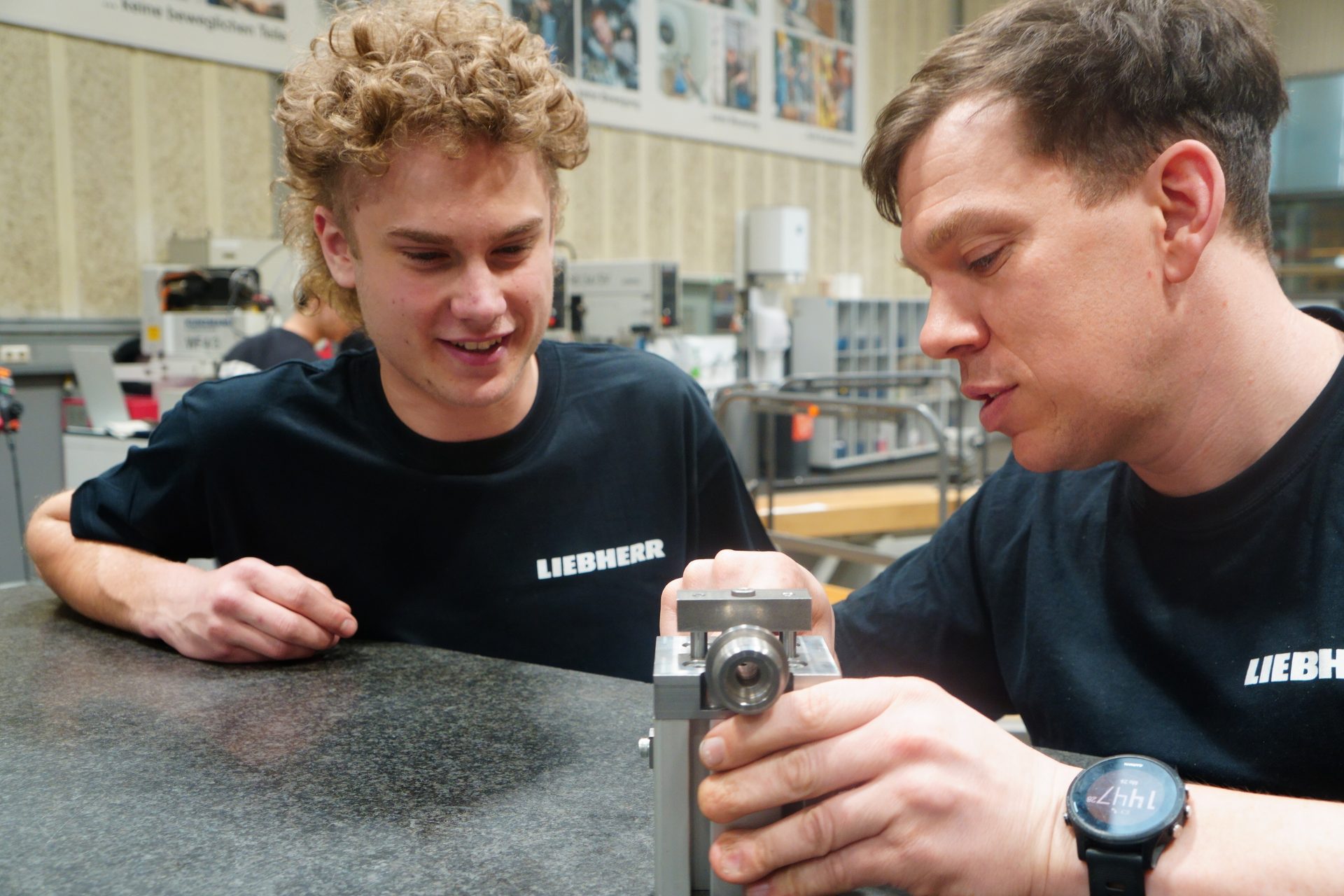 Two men, one with curly hair, in Liebherr shirts examining a small mechanical part on a table.