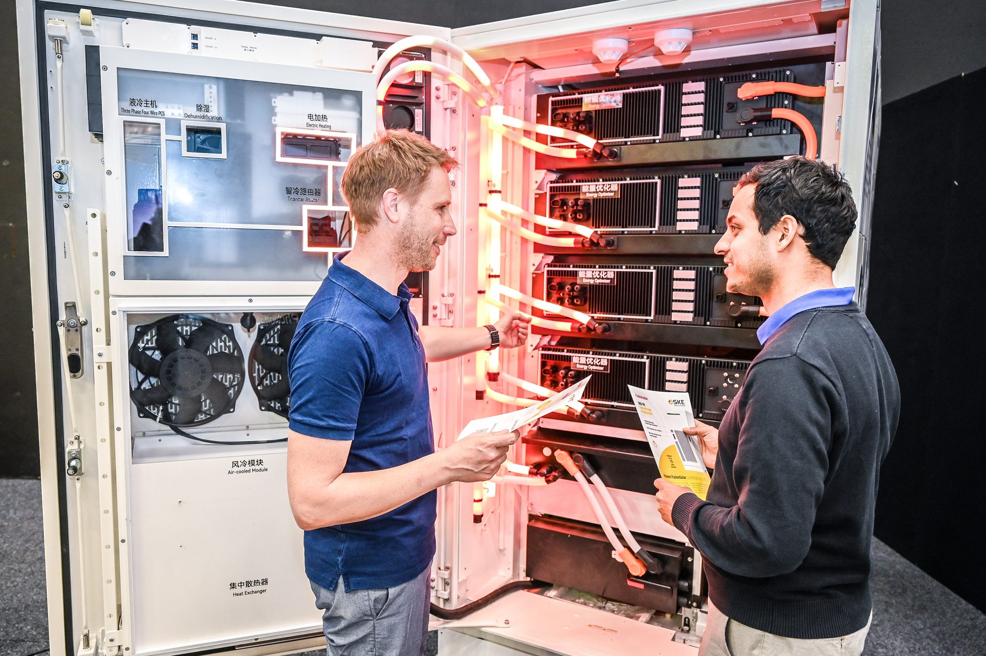 Two men discuss an intricate tech cabinet with glowing red tubes and internal components.