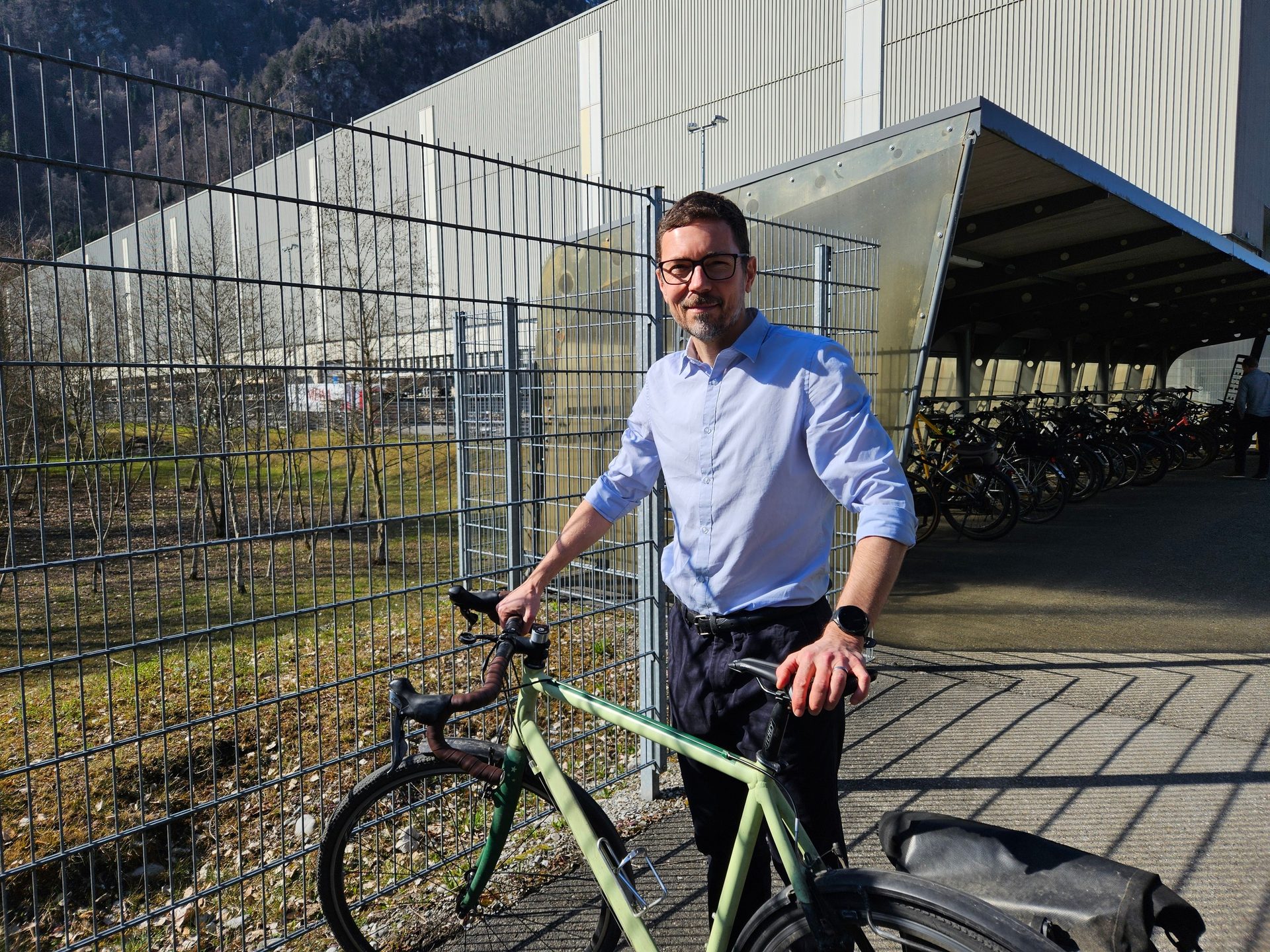 A man in a blue shirt and glasses stands smiling beside a light green bicycle, with a building and bike racks in the background.