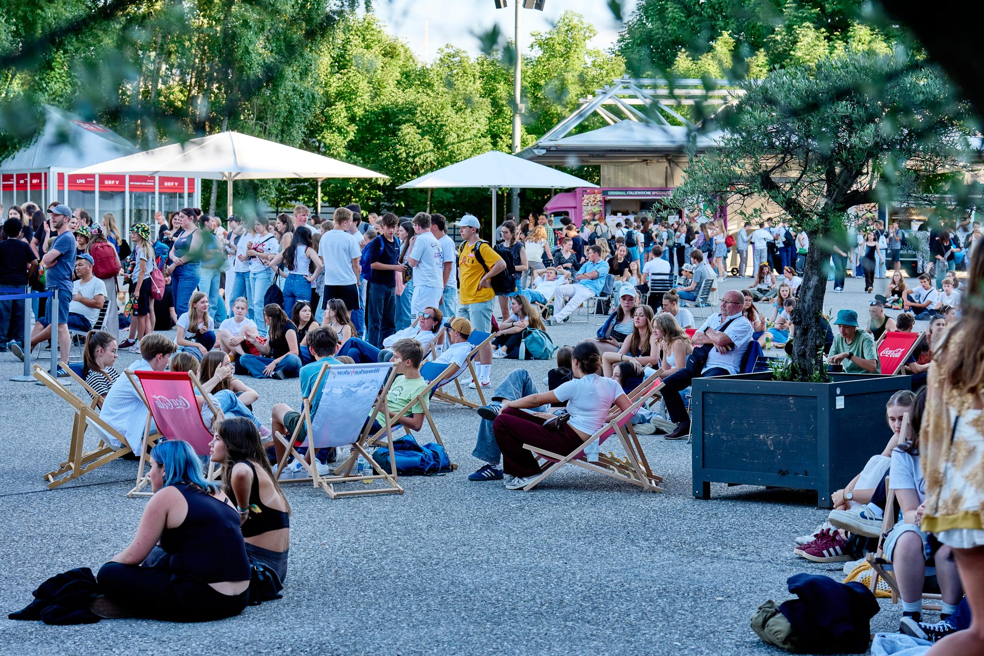 A large crowd of people socializing at an outdoor event with umbrellas, trees, and food stalls.