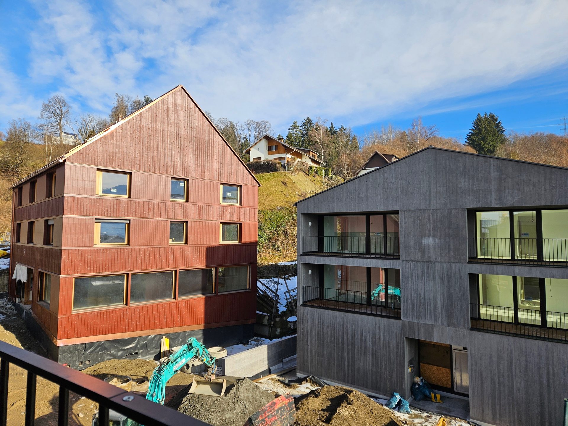 Two modern buildings under construction, one red-brown and one gray, with an excavator on site.