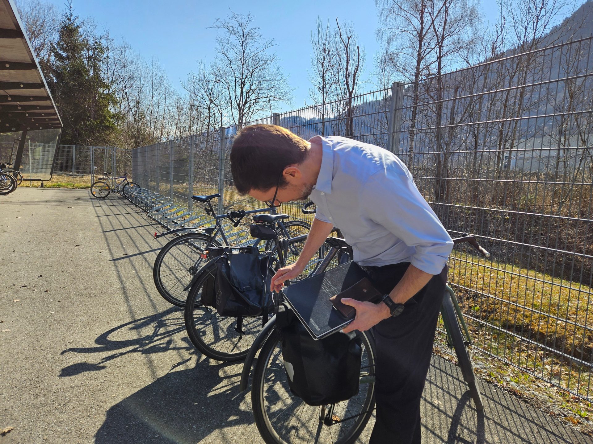 A man in a light blue shirt is interacting with a laptop and a bicycle parked at a bike rack on a sunny day.