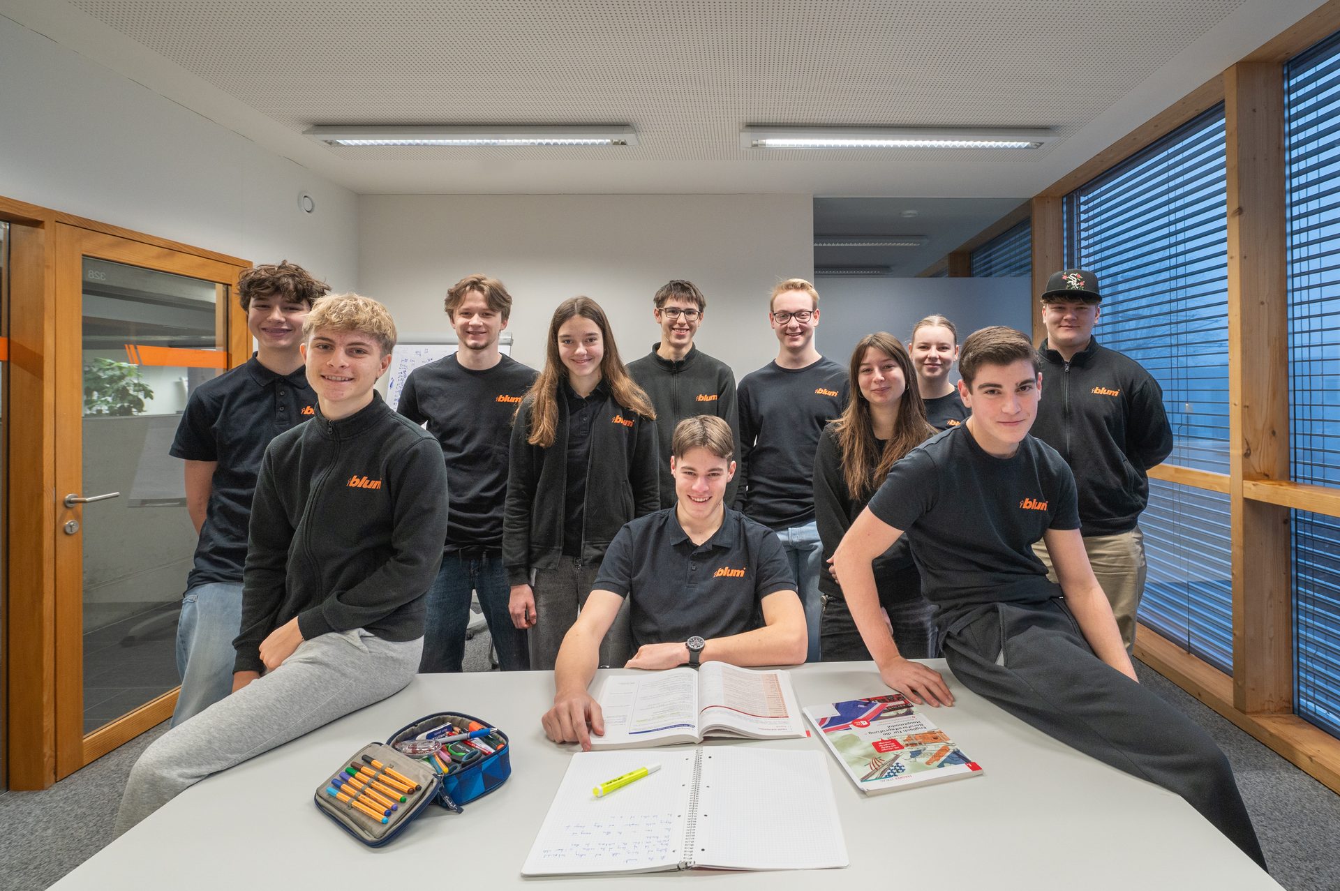 Ten young people in black "blum" branded tops pose in a modern office/classroom.