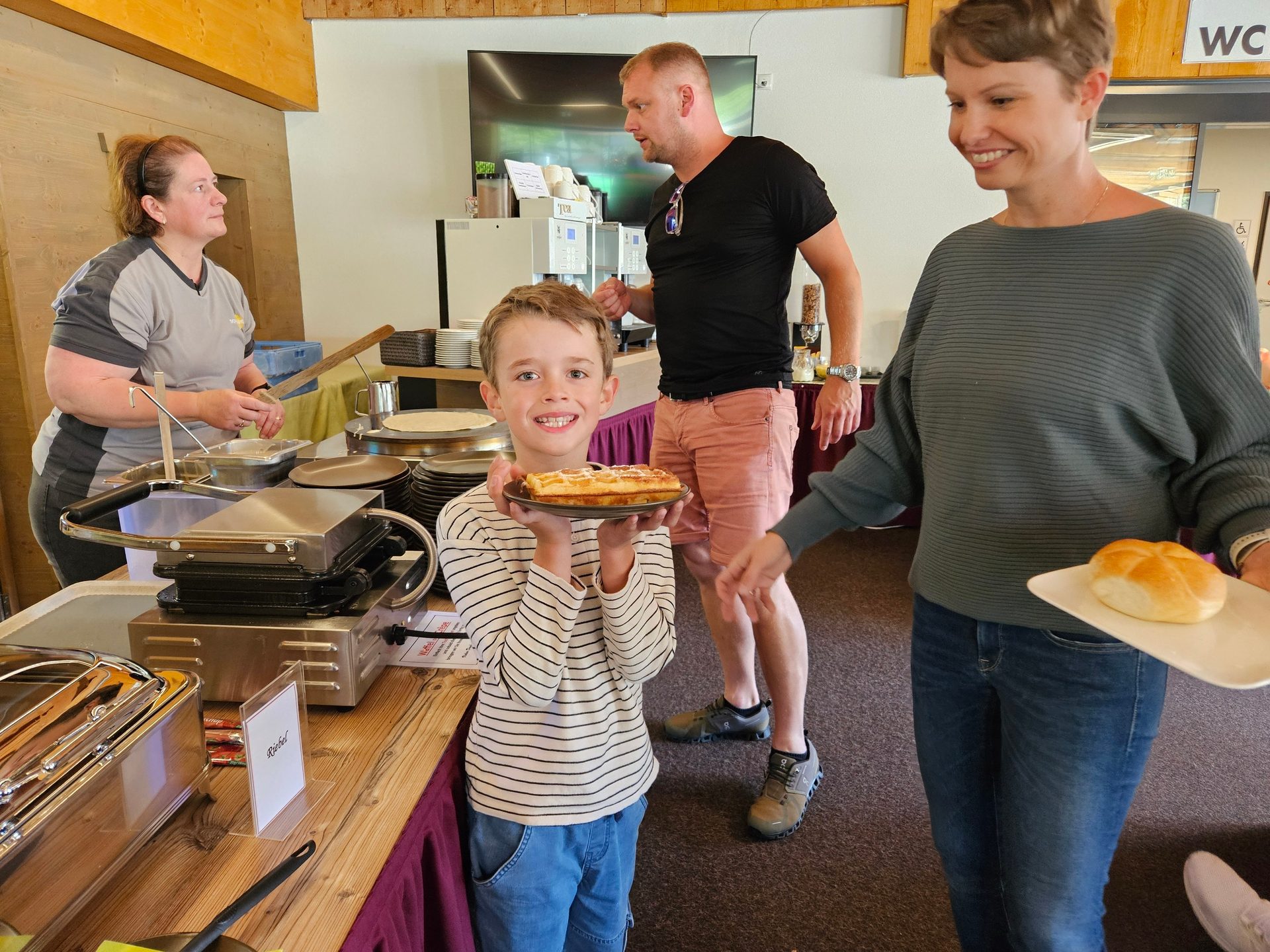 A boy holding a waffle, a woman with bread, and another woman making pancakes at a breakfast buffet.