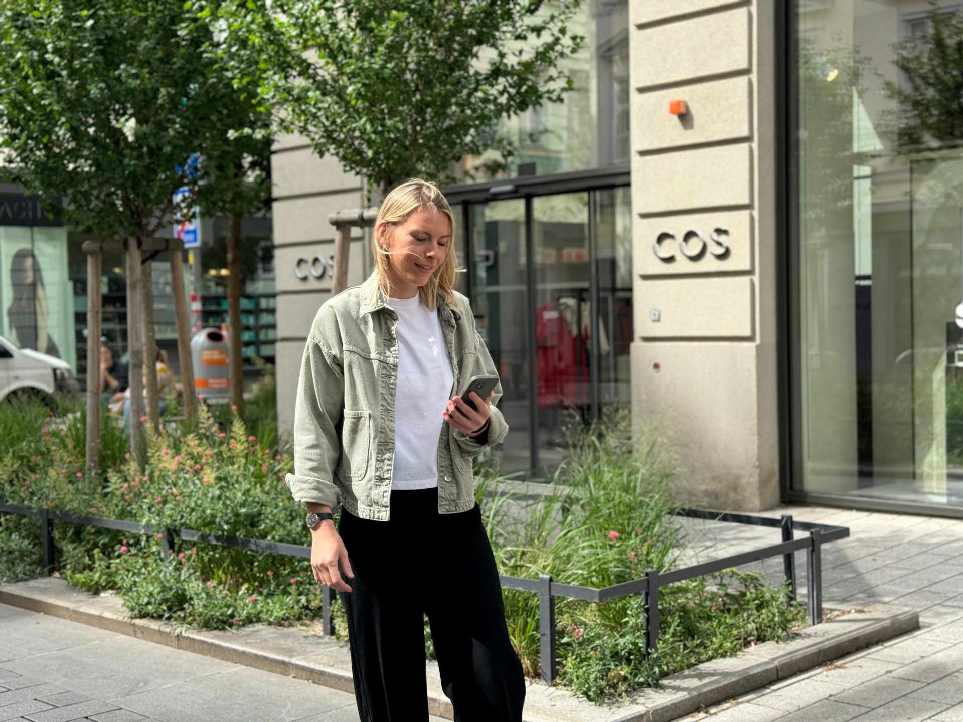A woman in a green jacket looks at her phone on a city street by a COS store.