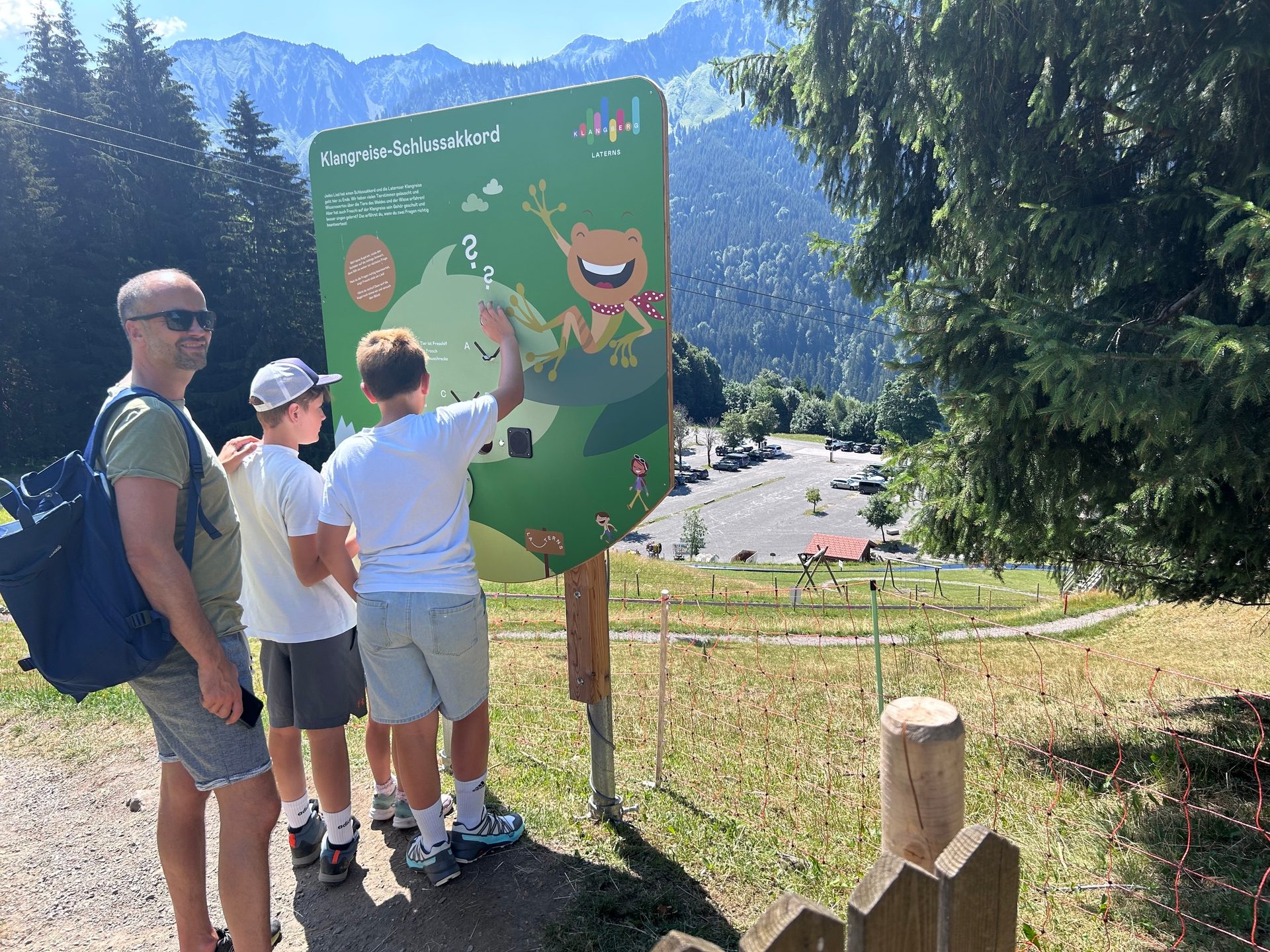 A man and two boys look at an interactive sign with a frog in the mountains.