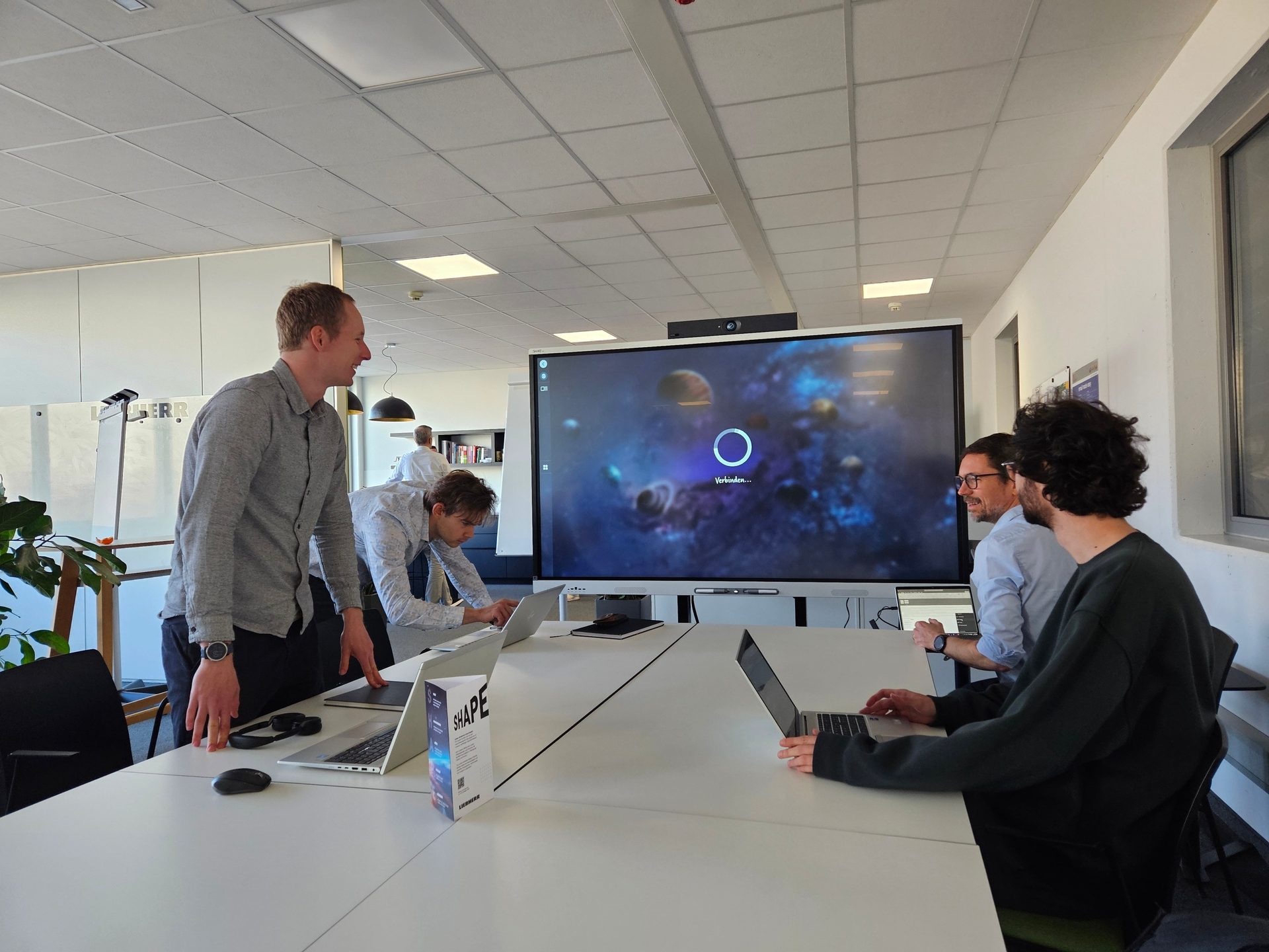Four men working together in an office with laptops and a large screen showing a cosmic scene.