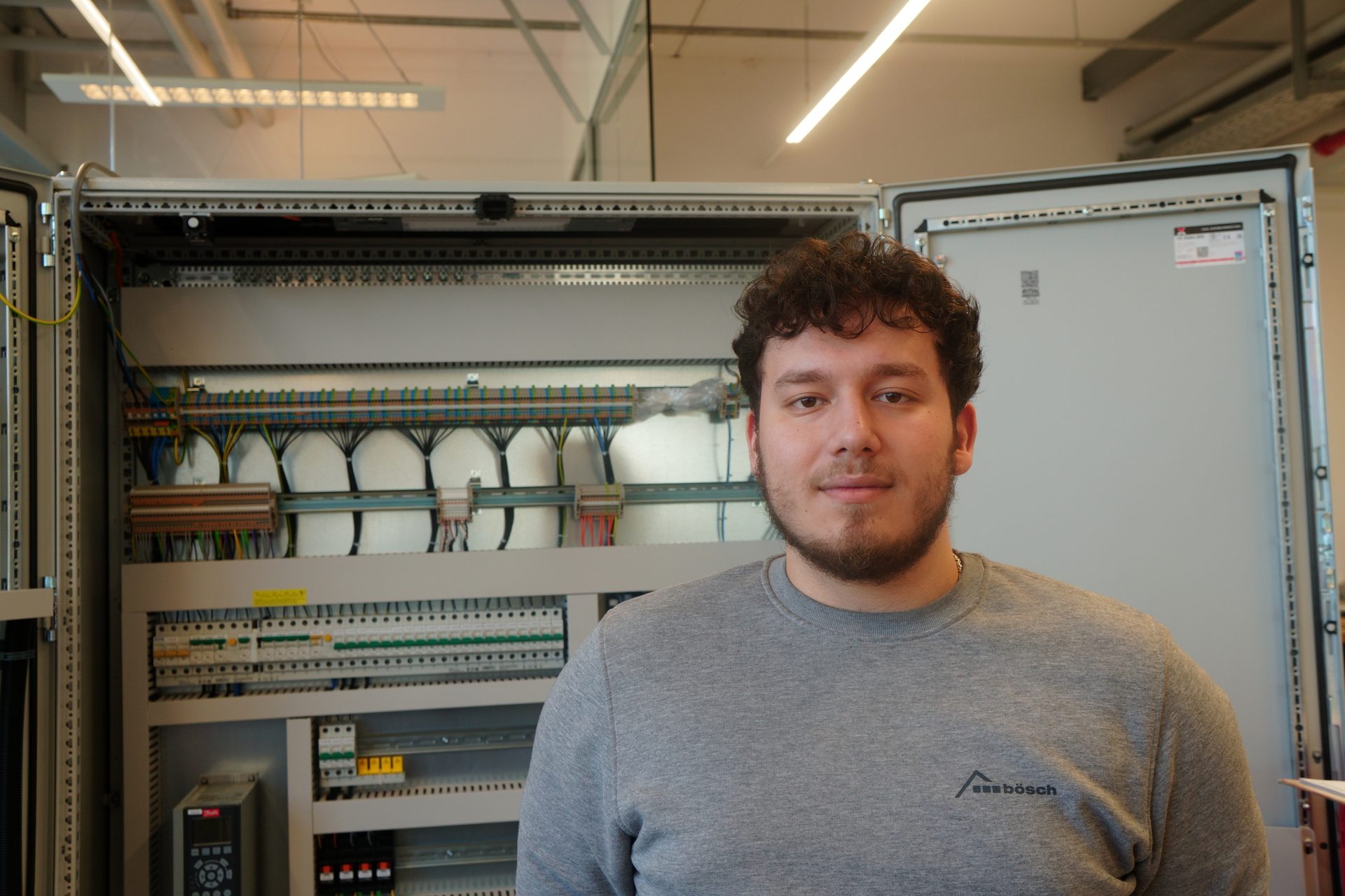 Young man with beard in front of an open electrical control panel with wires.