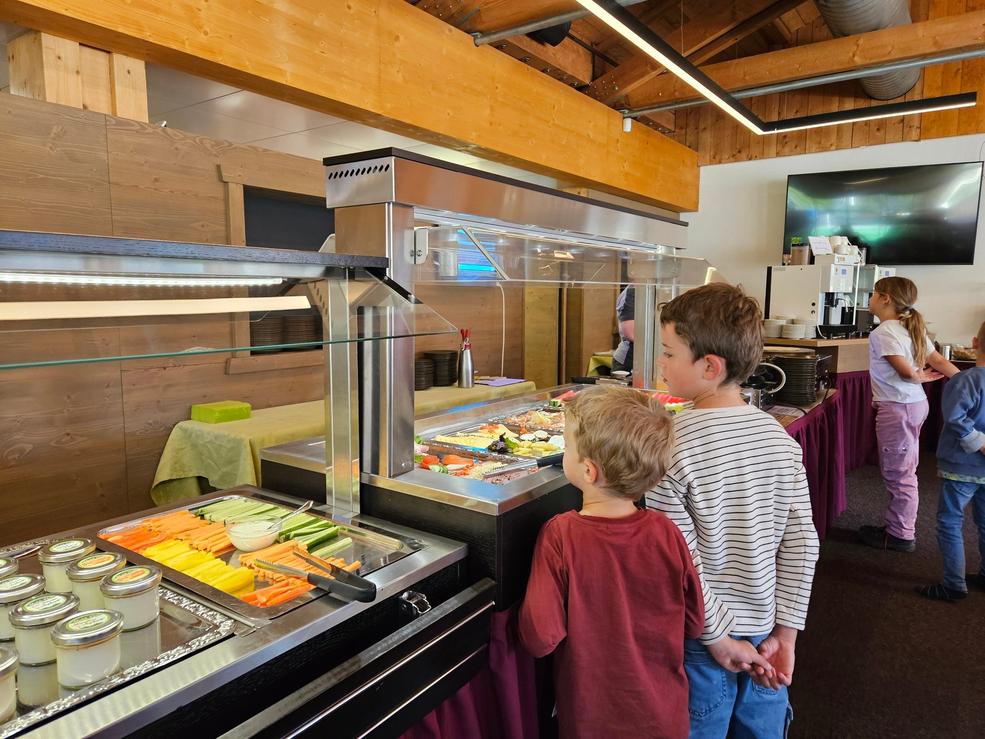 Children selecting food from a self-service buffet line with fresh vegetables, yogurt, and hot dishes.