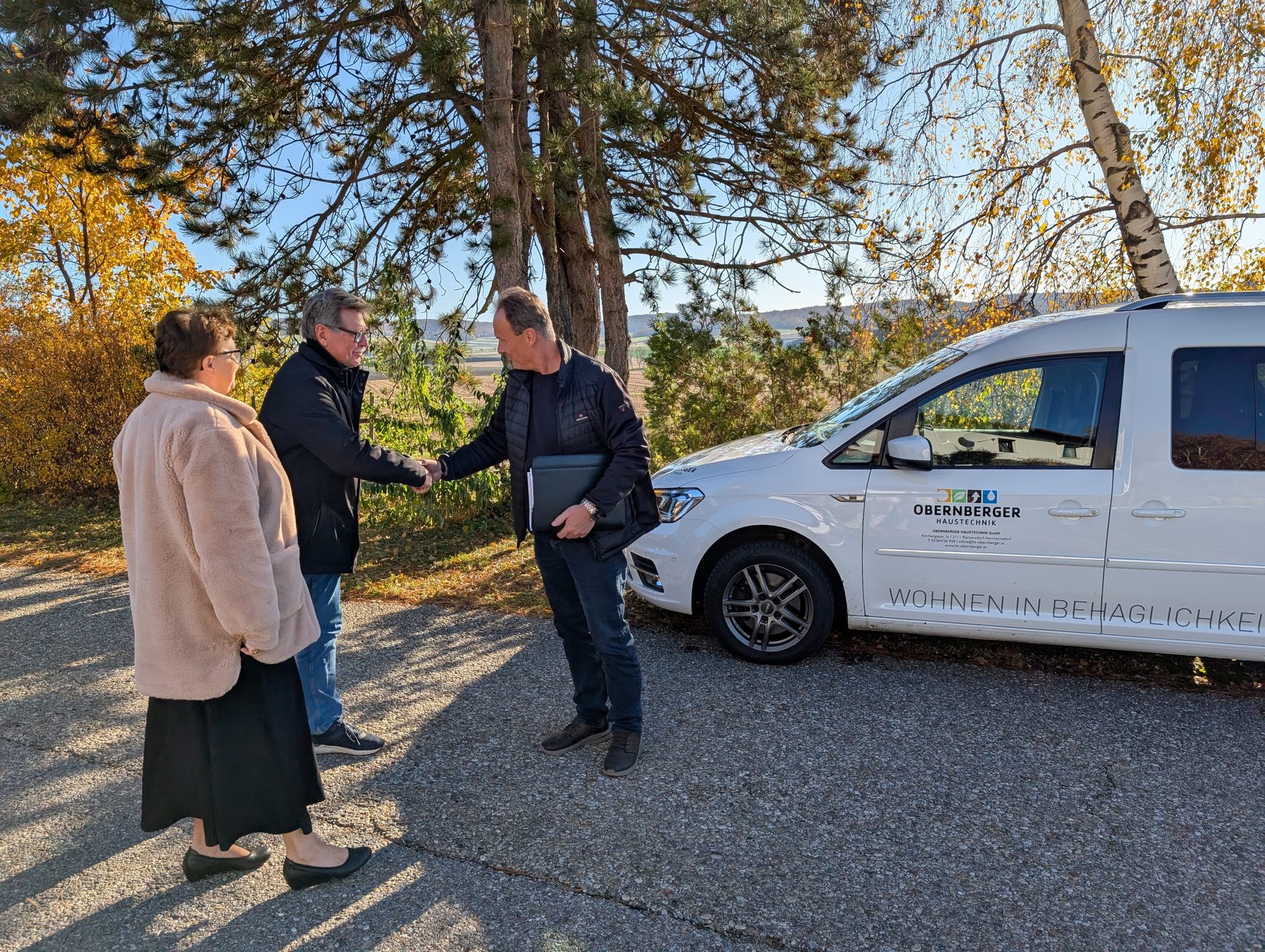 Two men shaking hands, a woman observing, next to a white van with "OBERNBERGER HAUSTECHNIK" and "WOHNEN IN BEHAGLICHKEIT" printed on it, surrounded by trees and a field.