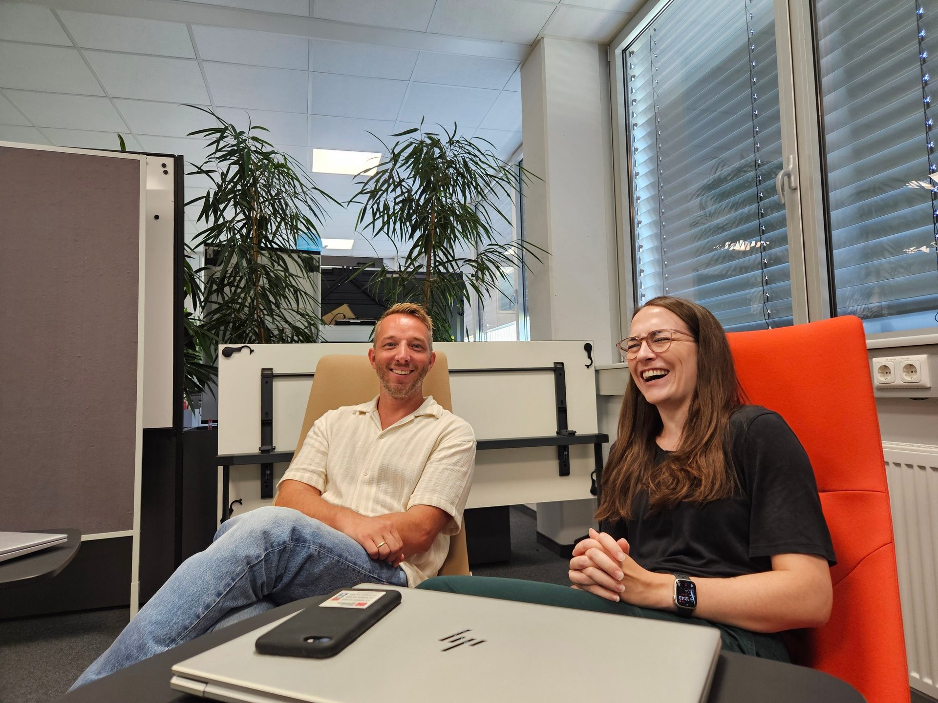 Two laughing colleagues, a man and a woman, seated in an office.