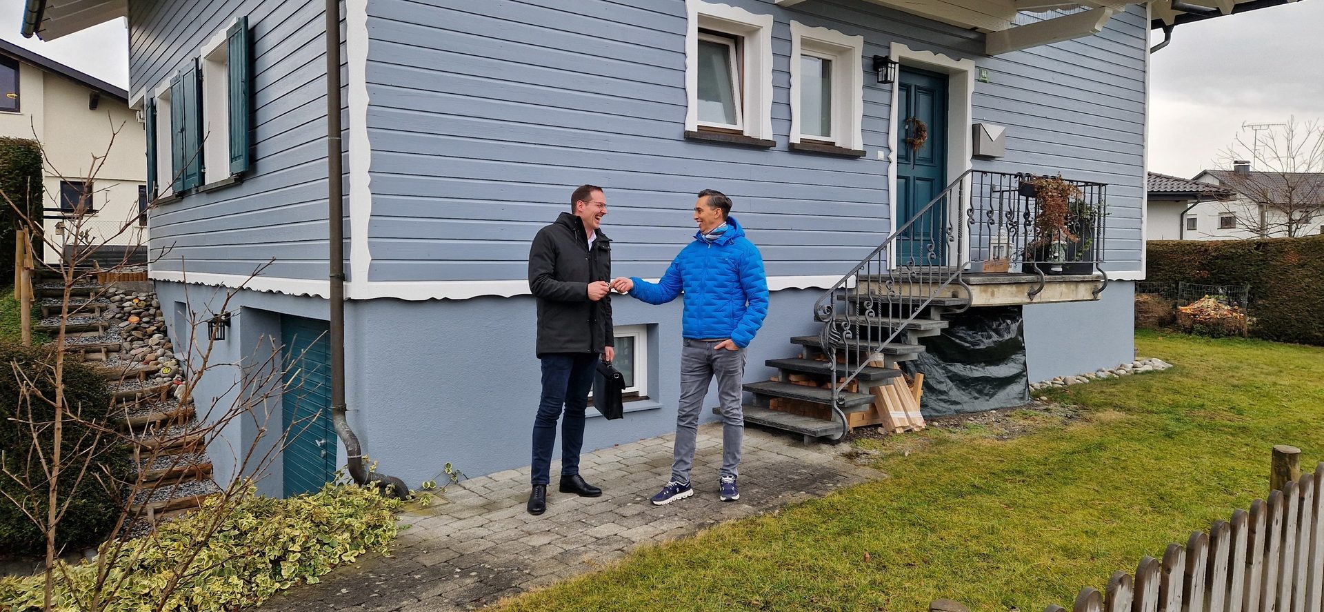 Two smiling men fist-bumping in front of a blue house, one in a dark coat, the other in a blue jacket.