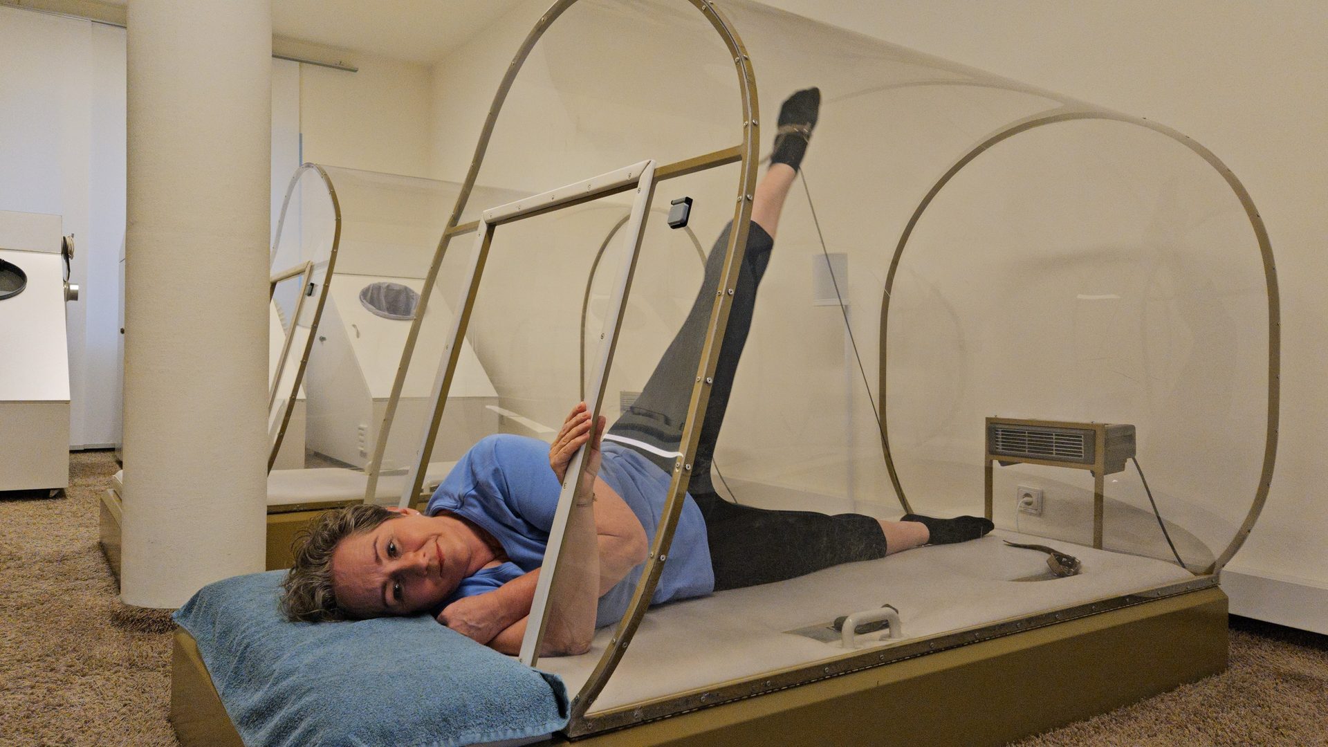 Woman in a clear chamber, lying with one leg raised high, head on a pillow looking at camera.