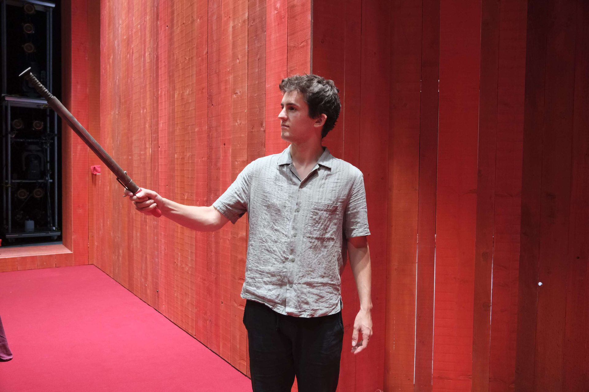 A young man in a grey shirt holds a long rod on a red stage.