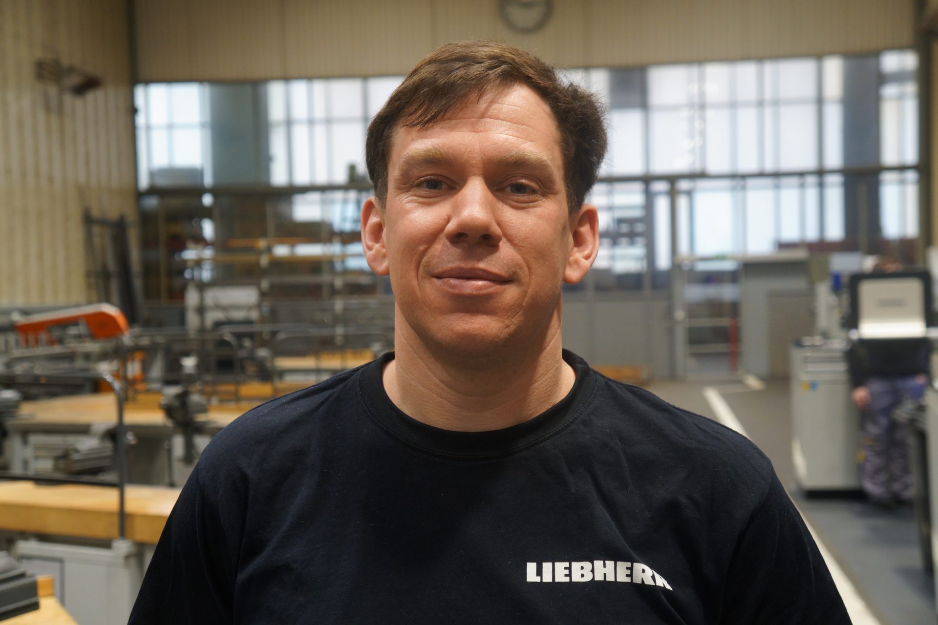 A man wearing a black Liebherr t-shirt looks directly at the camera, standing in a workshop.