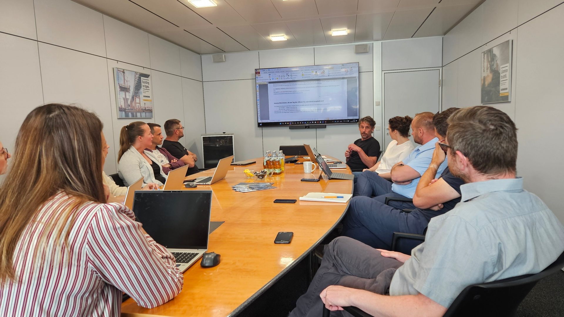 A business meeting with several people around a table, a screen shows a presentation.
