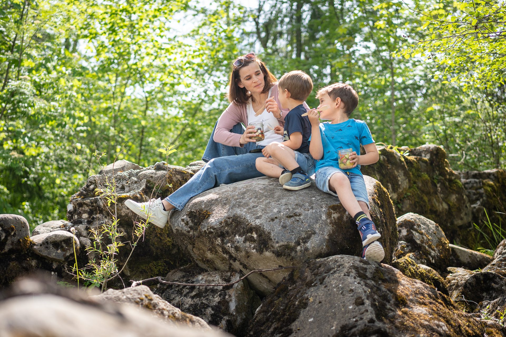 Mother and two young boys enjoying a picnic with fruit and granola while sitting on rocks outdoors.