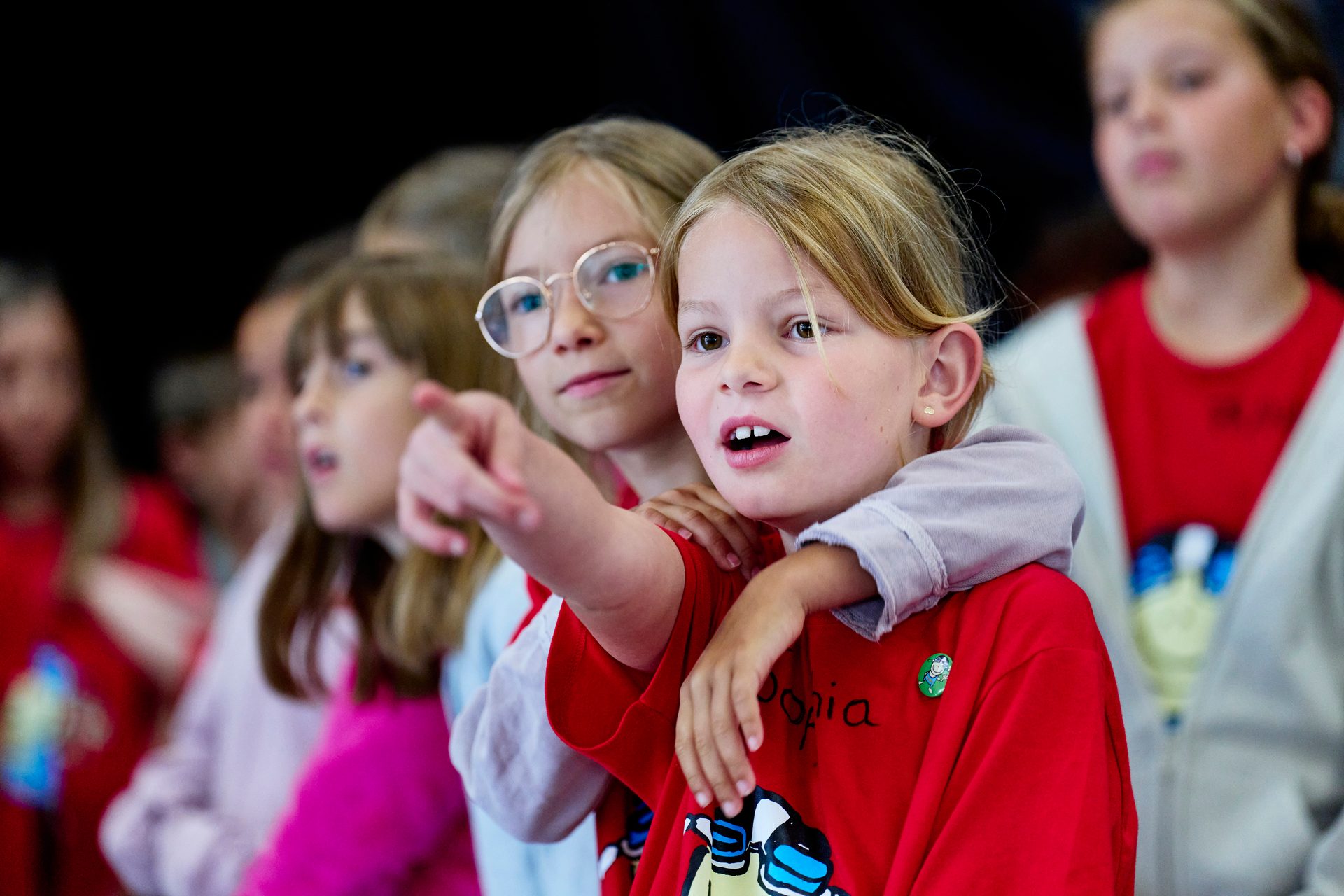 Two young girls, one pointing, look intently at something during an event, surrounded by other children.