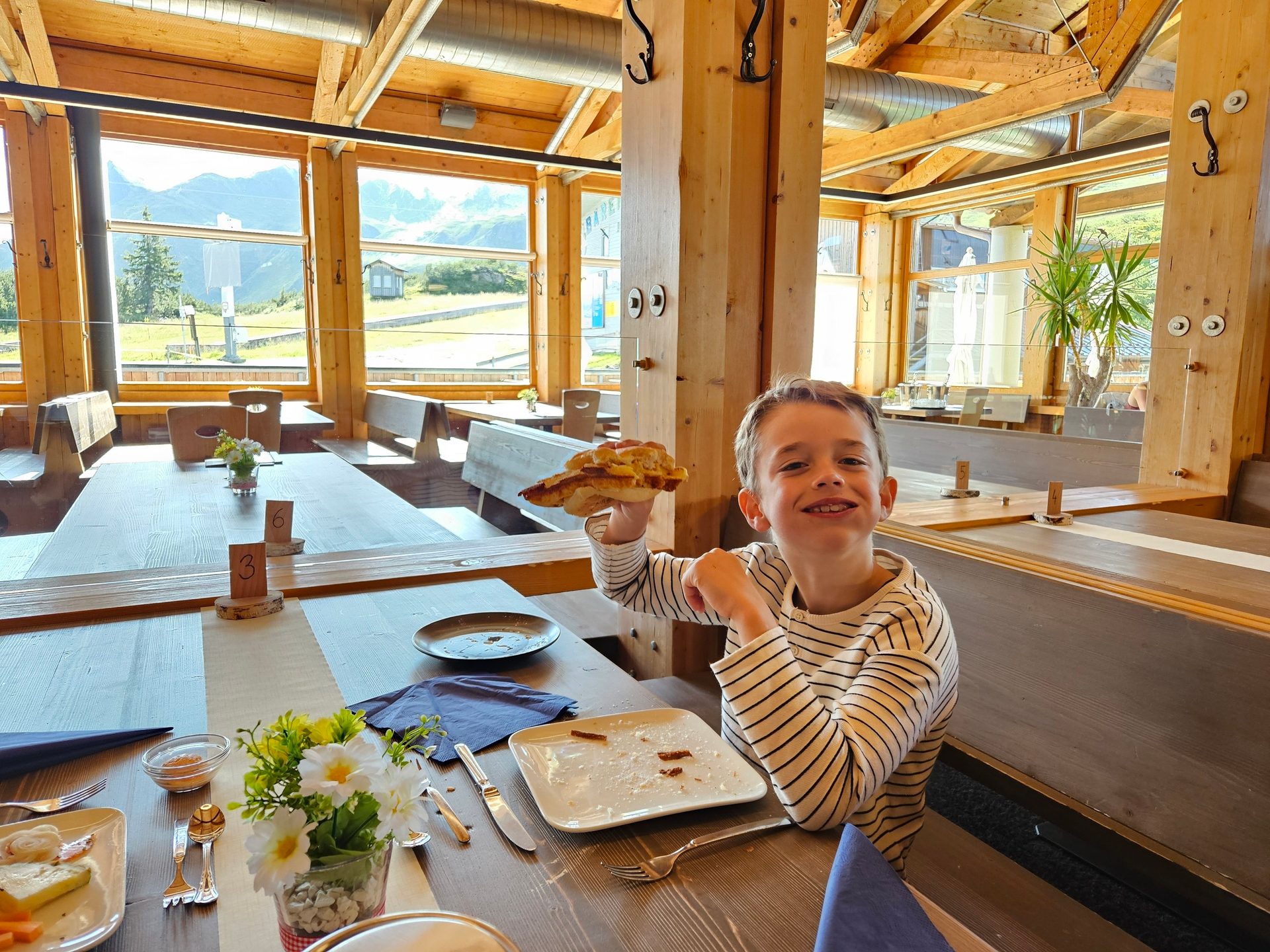 A boy smiles holding food in a rustic wooden restaurant with large windows overlooking a scenic mountain view.