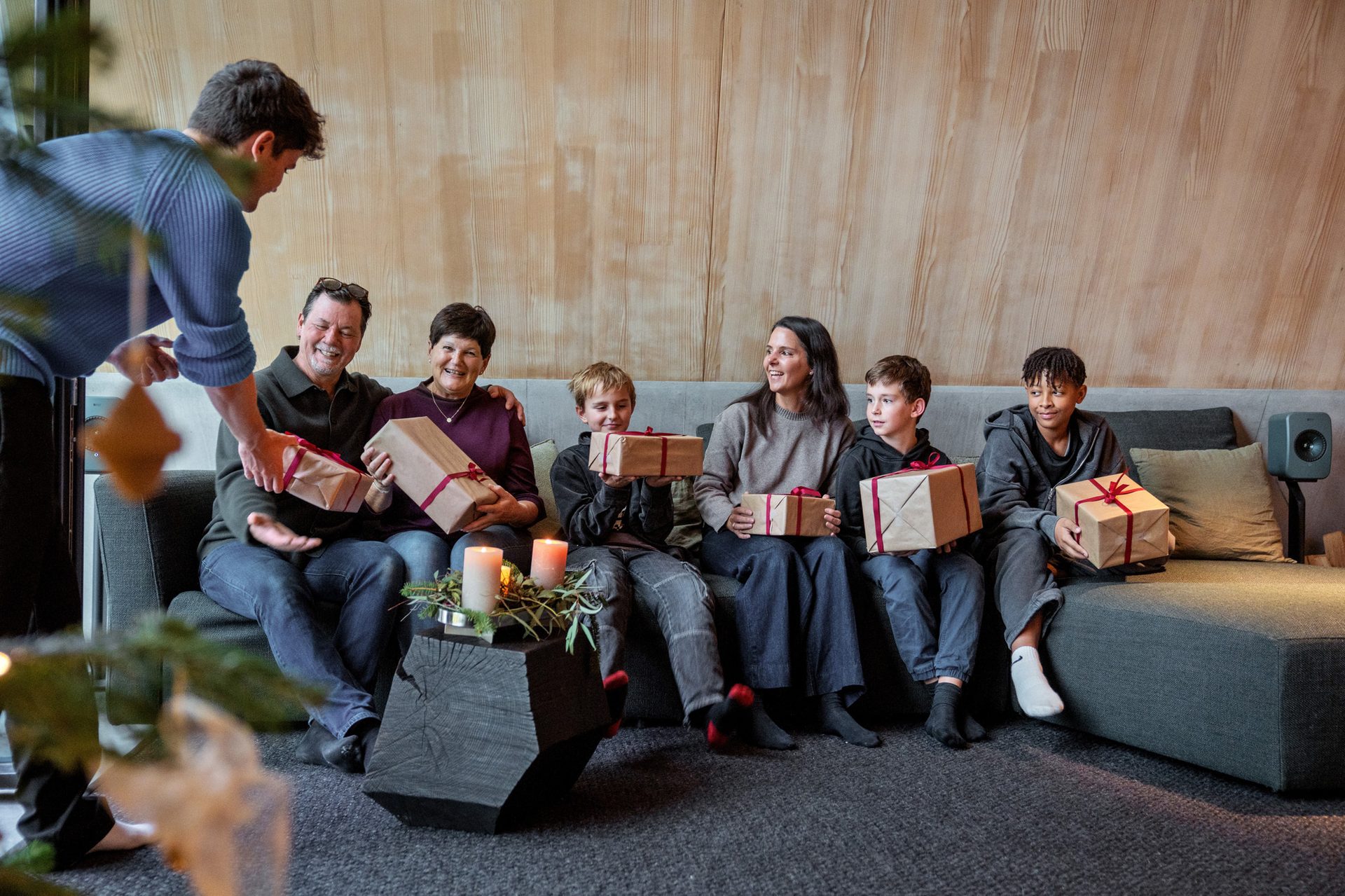 A family on a couch smiles, receiving presents from a standing person.