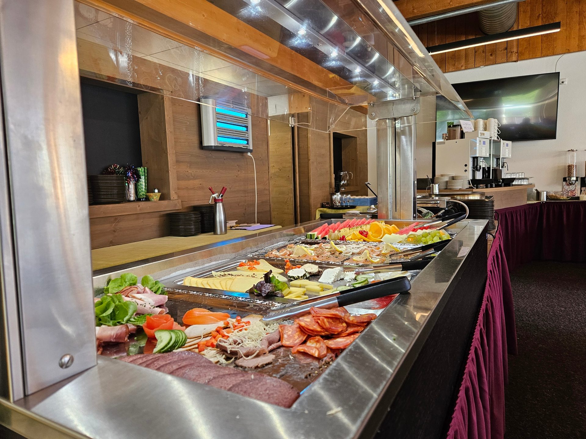 Self-serve cold buffet with assorted meats, cheeses, and fruits on a stainless steel counter.