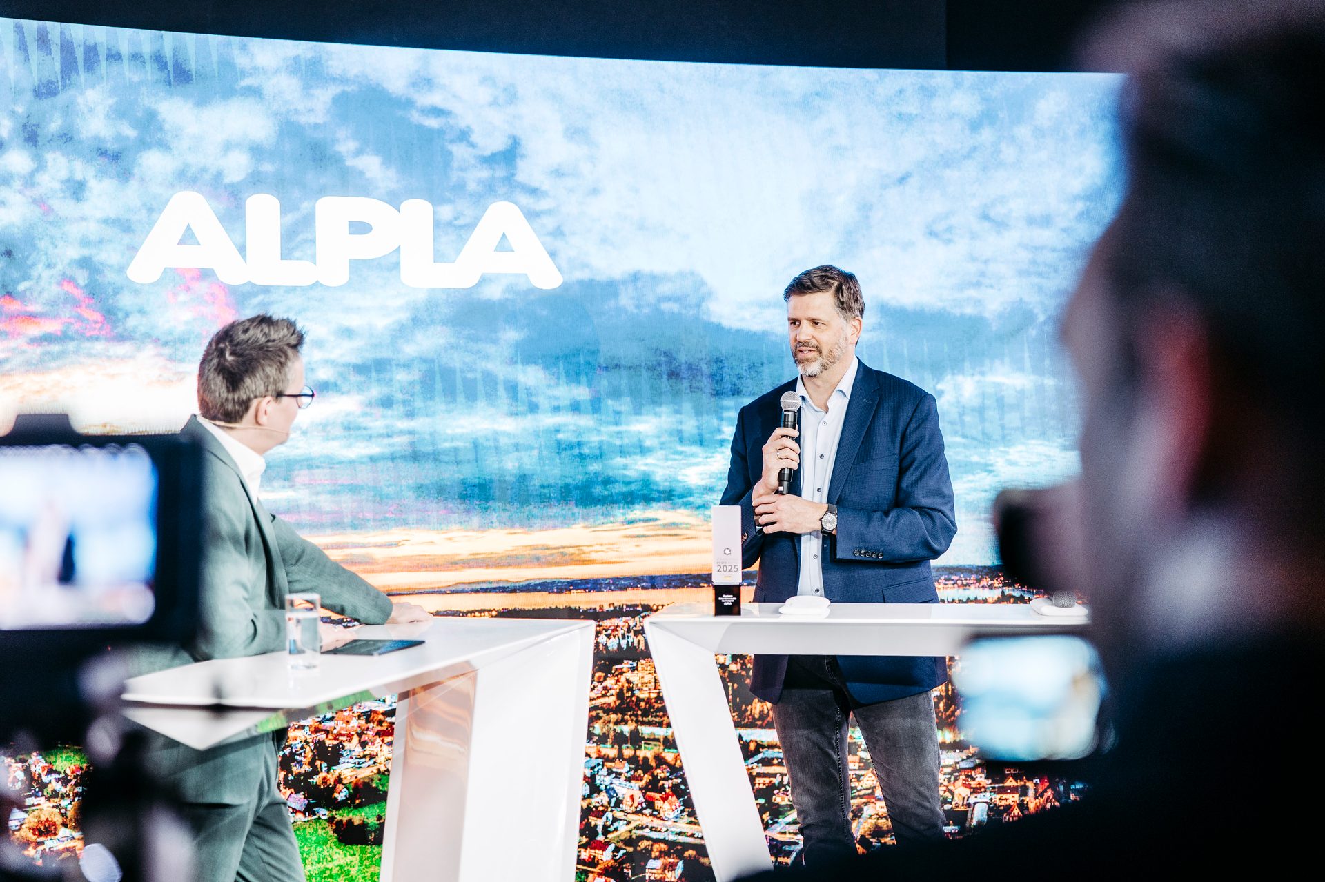 Two men on a stage with a large screen displaying the "ALPLA" logo and a landscape behind them.