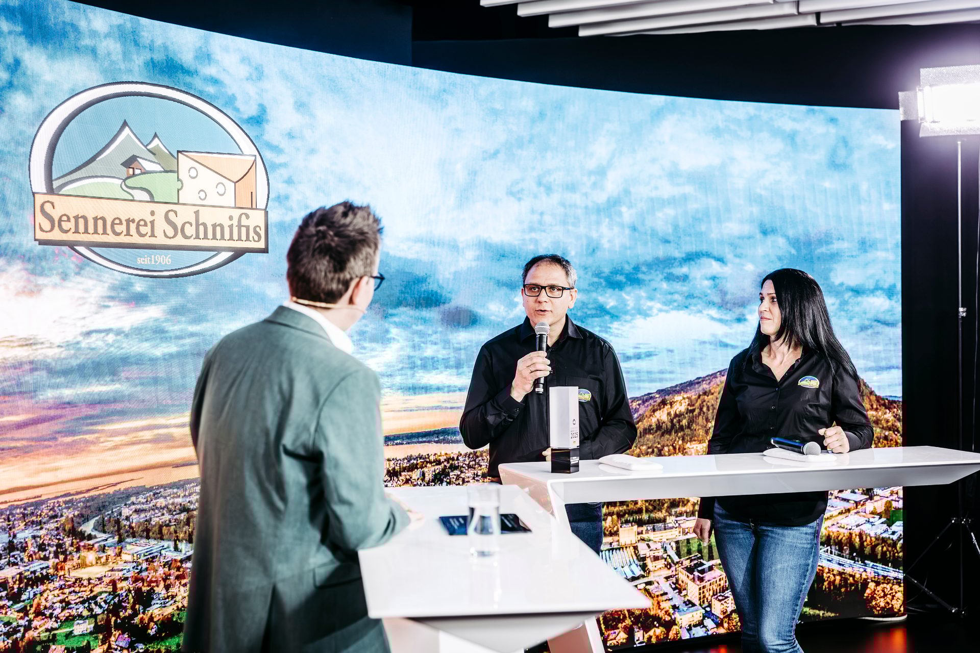 Three people, two presenting at a white table, one with a microphone, in front of a screen with a "Sennerei Schnifis" logo.